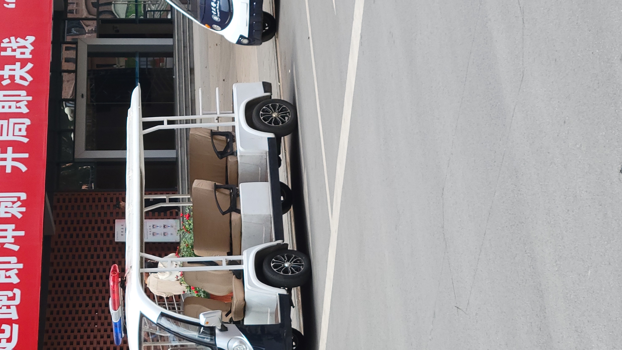 The image shows a vehicle parked on the street in front of a brick building with a red banner. The vehicle is white and black, with multiple rows of seats. There is a sign on the building. Another vehicle is partially visible in the background. The exact city is unknown.