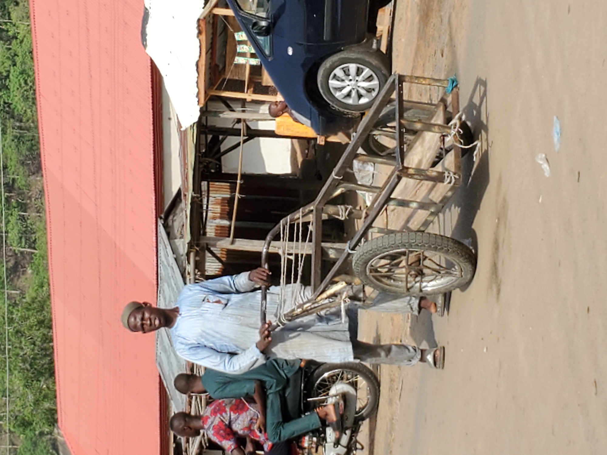 This media file captures an outdoor scene, likely a street or marketplace, in Mararaba, Nigeria, under clear daylight conditions. The image features several subjects and objects indicative of daily life.
In the foreground, a man stands centrally, looking towards the viewer. He wears a light-colored, long-sleeved traditional outfit, possibly light blue or off-white, and a matching cap. He is holding onto a large, unpainted metal pushcart, which features a single wheel and a sturdy frame, suggesting its use for transporting goods. Ropes are visible tied to the cart.
Behind and to the left of the main subject, two other men are partially visible. One, wearing a dark green shirt, is seated, with only his upper body and head showing. Further to the left and slightly behind him, another man in a red and white patterned shirt is also seated. A motorcycle's rear wheel and frame are visible beneath them.
The background consists of various elements. On the left side of the frame, a building with a corrugated red roof stretches horizontally, behind which lush green vegetation suggests a natural landscape. On the right, the ground is unpaved, appearing dusty or sandy, with scattered plastic debris. In the mid-ground, a blue sedan car is parked, partially obscured by makeshift structures made of wood and corrugated metal, indicating simple buildings or stalls. Another person, possibly a child, in an orange garment, is visible near the blue car.
The overall scene conveys a candid snapshot of everyday activity, with people seemingly engaged in or pausing from their routines, amidst local architecture and modes of transport. No discernible text is visible in the image.