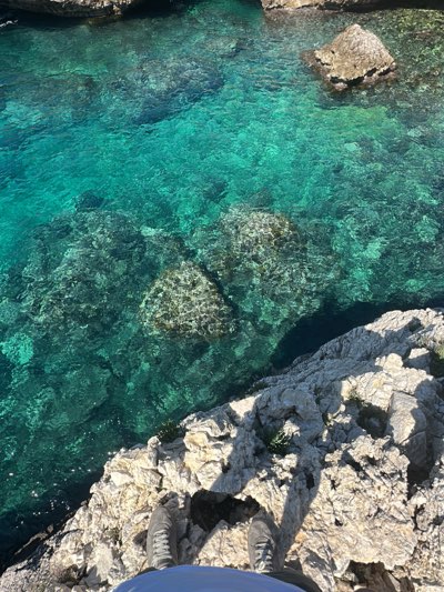 A top-down, eye-level shot shows someone standing on rocky terrain at the edge of clear, turquoise water in Nardò, Italy. The person is partially visible from the waist down, wearing a blue shirt or shorts and grey sneakers. The water reveals submerged rocks and pebbles, creating an intricate, textured pattern. Larger rocks are visible above the water in the background. The rocky coastline is light in color, contrasting with the vibrant water.