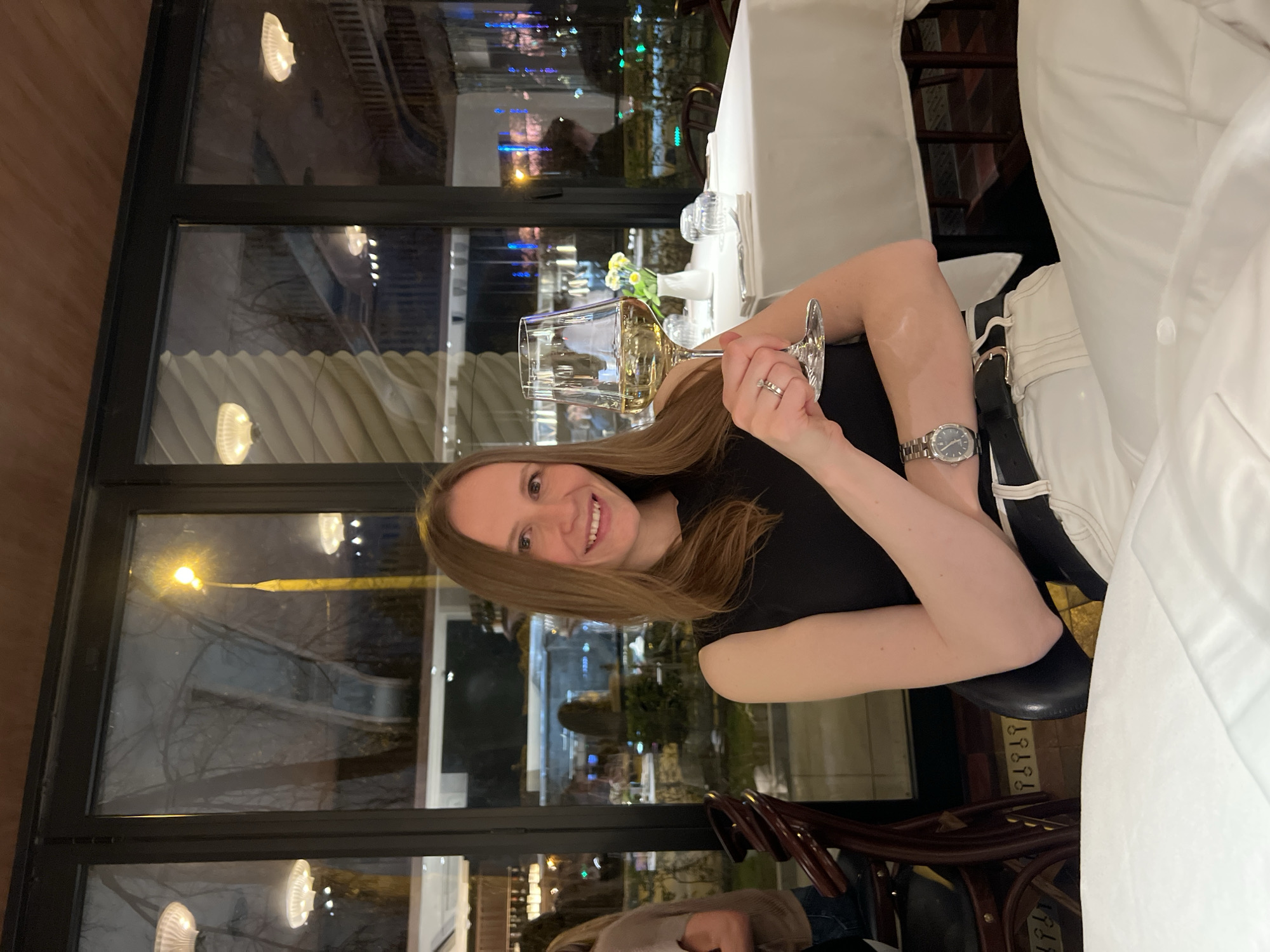 A smiling woman with long light brown hair is captured in a portrait shot, seated indoors at what appears to be a restaurant or upscale dining establishment. She is wearing a black top or dress and holds a wine glass filled with a light-colored beverage, likely white wine or champagne, in her left hand. A silver-toned watch with a dark face adorns her left wrist, and a ring is visible on her left ring finger.
The setting indicates it is nighttime, with the background dominated by large windows framed in black, looking out onto a dark exterior illuminated by streetlights and the distant glow of city lights and reflections. Trees are visible through the window panes. Inside, the reflection of interior lighting fixtures, some with a distinct shell-like shape, can be seen in the glass.
To the woman's right, a table is set with a crisp white tablecloth, additional glasses, and a small vase holding yellow flowers. Part of a dark wooden chair is visible in the foreground. The woman's direct gaze and bright smile convey a sense of happiness and enjoyment, suggesting she is having a pleasant evening out. No specific text is visible in the image.