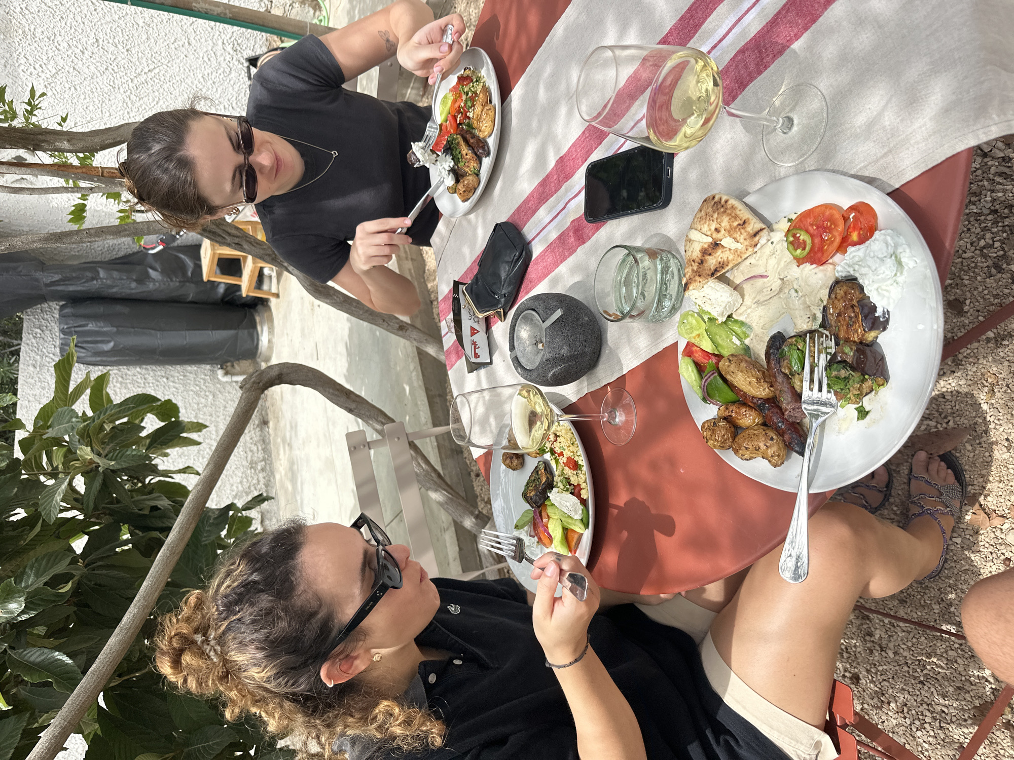 Two women are seated at a table outdoors, eating lunch. One woman wears sunglasses and a black t-shirt. The other woman is wearing glasses, a black polo shirt, and light shorts. A partially consumed lunch sits on each of their plates. The table is set with a white tablecloth with red stripes. On the table are two glasses of white wine, a water glass, a cell phone, a black wallet, and a small stone ashtray. A tree branch hangs over the lower left of the frame. The location is not identifiable from the image.