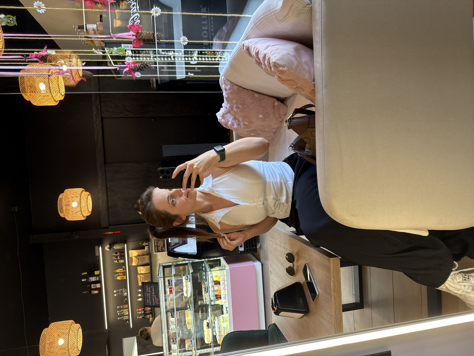 The image shows a woman taking a selfie in a cafe. She is sitting on a couch, wearing a white V-neck top and black pants. She has her hair in a ponytail and is holding her phone in her left hand. There are two pink pillows on the couch next to her. A brown handbag is also visible on the couch.
In the background, there is a display case filled with pastries and drinks. Behind the display case, there are shelves stocked with bottles. There are two woven lamps hanging from the ceiling, casting a warm glow. The scene appears to be indoors, possibly in a cafe or bakery, given the display of food and drinks. The reflection in the mirror shows a table with sunglasses and a phone on it. The overall atmosphere seems relaxed and casual.
There is some visible text in the background, but it is mostly illegible or too small to read clearly. However, one sign in the reflection appears to have the word "HÖLLER" on it, possibly indicating the name of the establishment. The image suggests it is daytime, based on the lighting. The woman's expression is neutral, as she focuses on taking the selfie. The location context of Szklarska Poręba, Poland, is provided, suggesting the cafe is situated in this town.