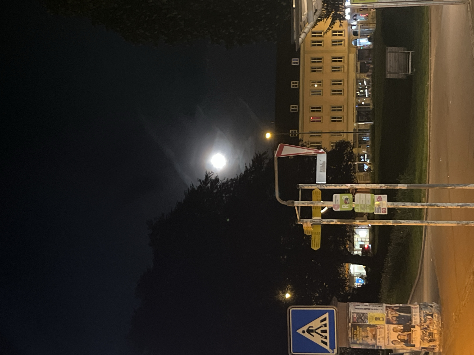The image depicts a night scene in an urban environment. The moon is visible in the sky, partially obscured by clouds. In the foreground, there is a street with a crosswalk sign. Buildings can be seen in the background, illuminated by streetlights and interior lights. The scene is a street view with a focus on the intersection.