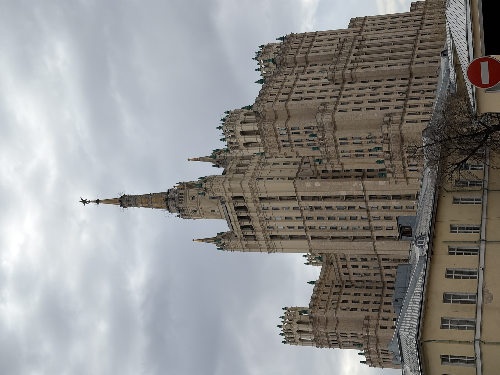The image depicts a tall, ornate building against a cloudy sky. The building is likely one of the Seven Sisters, Stalinist skyscrapers located in Moscow, Russia. The structure has a spire topped with a star. The building is light-colored with numerous windows. A street with a smaller building and a red and white "no entry" sign is visible in the foreground. The scene is urban and the weather is overcast.