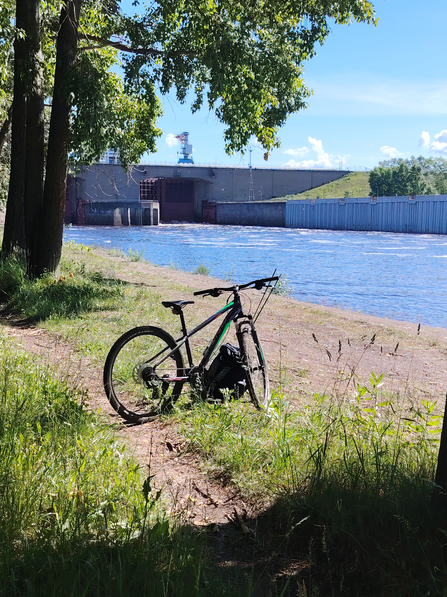 An outdoor, eye-level, medium shot shows a black and green bicycle parked on a grassy bank next to a wide, flowing river. The bicycle is positioned on the right side of the frame, angled slightly towards the river. Behind the bicycle, the river is a bright blue with whitecaps and ripples, suggesting a current. Further back, a large concrete dam structure stretches across the width of the image, with water flowing over its edge. On the far right, a tall, industrial crane with a blue boom is visible in the distance, with a plume of white smoke or steam rising from it. The left side of the frame is dominated by the dark trunks and green leaves of a tree, providing shade. The sky is a clear, bright blue with a few scattered white clouds. The image appears to have been taken on a sunny day. The scene is set at the Kama River dam in the city of Chaikovsky, Russia, a location known for its hydroelectric power station.