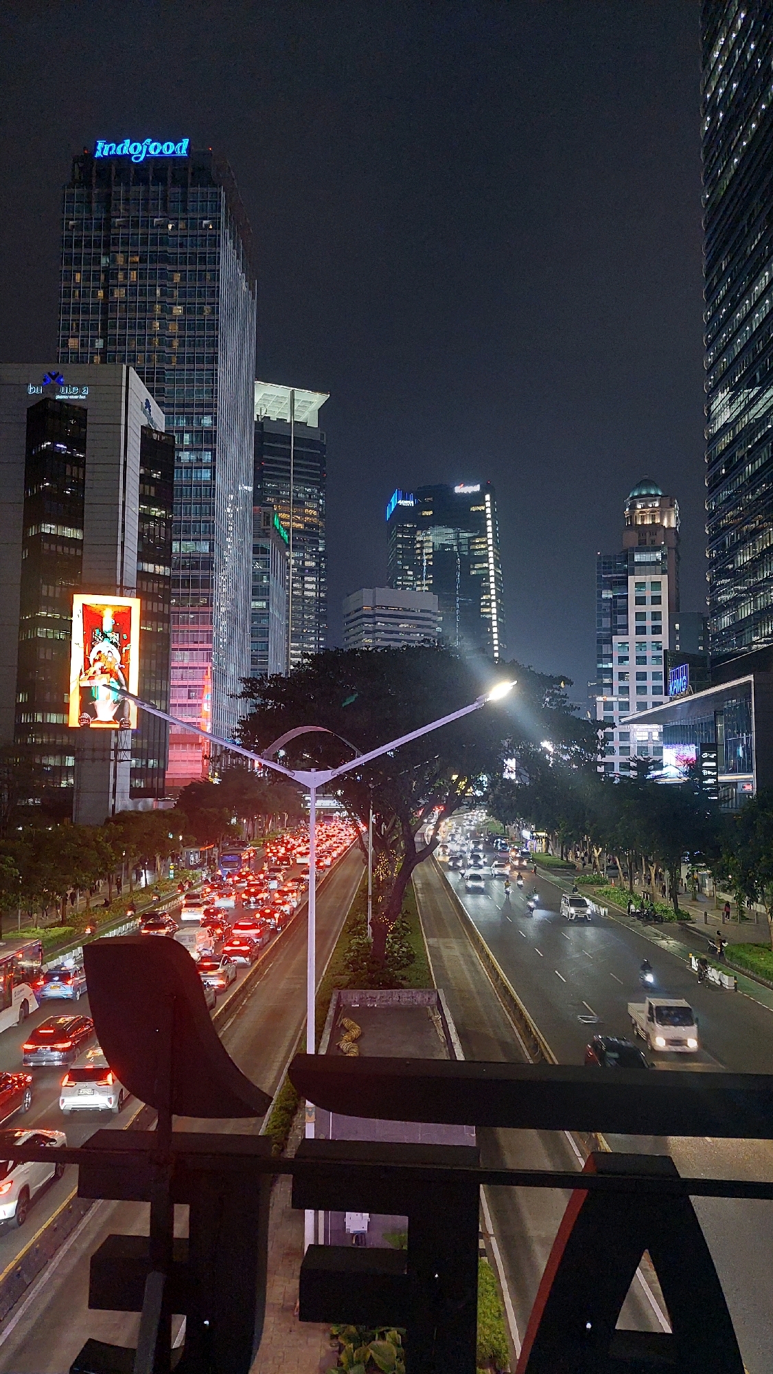 The image depicts a nighttime street scene in Central Jakarta, Indonesia, characterized by heavy traffic and illuminated skyscrapers. In the foreground, a dark, abstract metal sculpture partially obscures the view. Beyond the sculpture, a multi-lane road is congested with numerous vehicles, their headlights and taillights creating streaks of light. The road is lined with trees and streetlights, some of which are bent and ornate.
The urban environment is dominated by tall, modern buildings that are brightly lit, showcasing their architectural designs and window patterns. One prominent building displays the "Indofood" logo in blue neon lights at its apex. Another building features a large, illuminated advertisement screen with a red and orange graphic. The sky above is dark, indicating the late hour. Pedestrians can be seen on the sidewalks, and motorcycles navigate through the traffic. The overall atmosphere suggests a bustling metropolitan area at night.