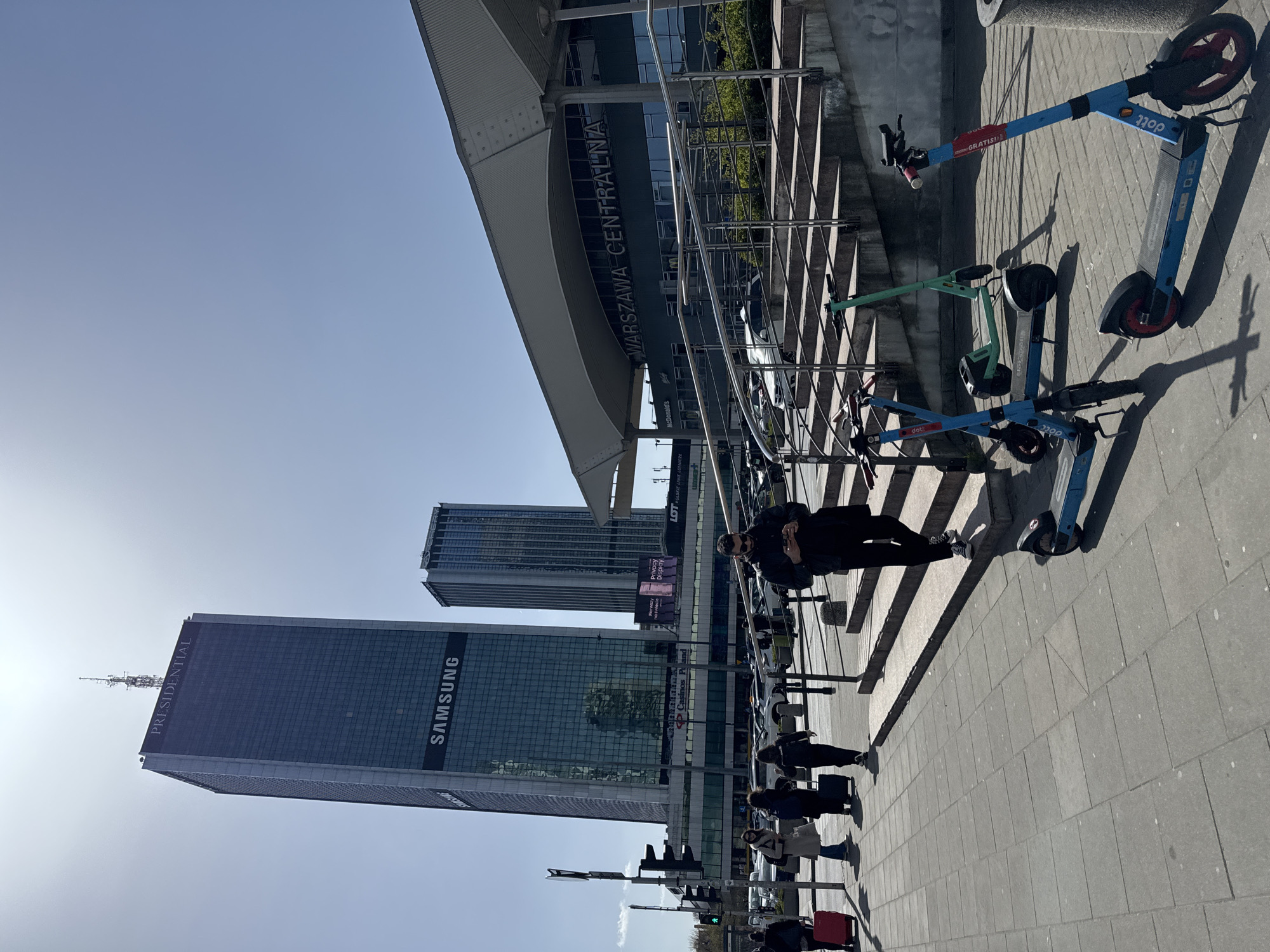 The image depicts a street scene in Warsaw, Poland, featuring modern architecture. In the foreground are a group of electric scooters parked near Warsaw Central Station ("WARSZAWA CENTRALNA" signage visible), and a number of people walk along the sidewalk. Two prominent skyscrapers rise in the background; one displays the "SAMSUNG" logo. Signage for other establishments such as McDonald’s and LOT (Polish Airlines) is visible. The sky is clear.