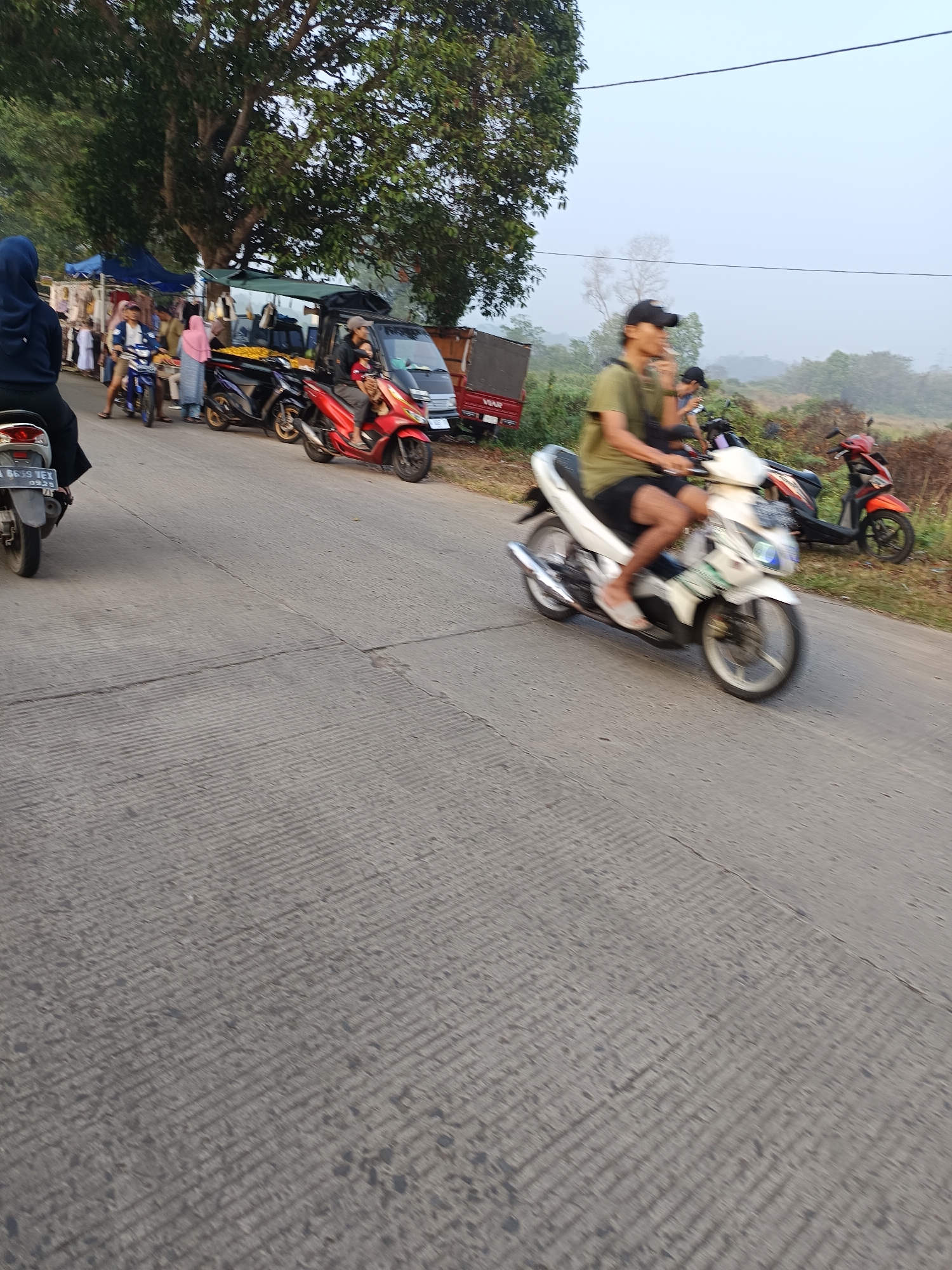 The image depicts a street scene in Sodong, Indonesia, likely during the daytime with a hazy sky. In the foreground, a person is riding a white motorcycle, captured in motion with a blurred effect. They are wearing a green t-shirt and dark shorts. To the left, a dark-colored motorcycle is parked, and further left, another person wearing a blue headscarf is seated on a motorcycle. In the middle ground, several stalls are set up along the side of the road, displaying various goods, and people are milling around them. Several other motorcycles, including a red one, are visible parked and in use. Behind the stalls, a small truck with a red canopy is parked. The environment is characterized by a paved road with textured markings, a large tree providing shade, and vegetation in the background.