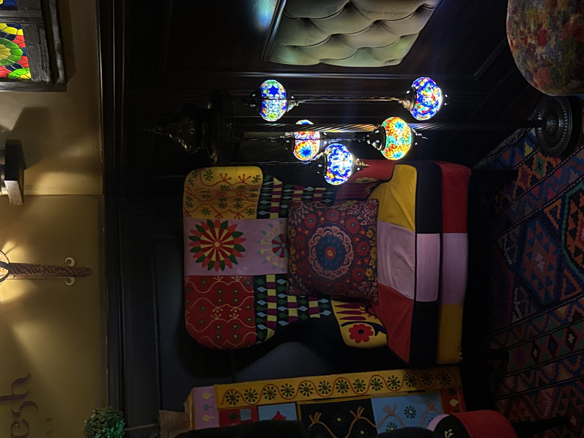 The scene depicts a dimly lit, ornate interior lounge area within an establishment in Yerevan, Armenia. A prominent wingback armchair, upholstered in a multi-paneled patchwork fabric featuring sections of red, yellow, black, and pink with various geometric and floral patterns, occupies the central left. A circular, intricately patterned cushion in red and blue rests on the armchair's seat. To the right of the armchair stands a dark-toned metal floor lamp, from which five illuminated, colorful mosaic glass globes are suspended, casting warm, variegated light.
The floor is covered by a patterned rug with geometric designs in shades of red, blue, and orange. The lower walls are lined with dark wood paneling, while the upper walls are painted a muted olive-gold. On the upper left wall, a decorative wall sconce is visible, along with a partial, stylized word, "esh." A rectangular stained glass panel featuring red, yellow, and green sections is mounted on the upper right wall. To the far right, a section of olive-green, tufted upholstered seating is visible. In the lower right foreground, a small, partially visible upholstered stool or ottoman with a floral pattern is present. On the far left, a tall, narrow decorative column or panel, adorned with colorful, stacked geometric patterns, stands next to a small potted topiary plant. No individuals are present, and no specific actions are being performed.