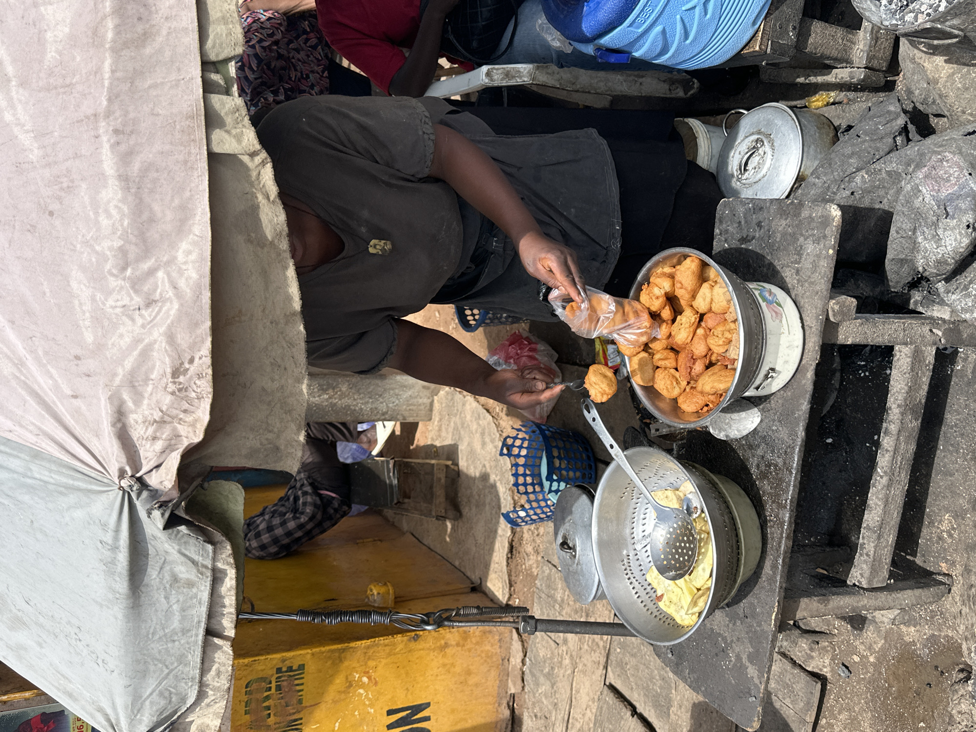 This image captures an outdoor street food stall in Gura Top, Nigeria, under bright daylight. The scene is viewed from a slightly elevated perspective, looking down at the vendor and their setup.
The main subject is a vendor, likely a woman, with dark skin, wearing a dark t-shirt and a dark apron or skirt. She is actively bagging fried food. Her right hand holds a clear plastic bag, while her left hand uses a metal slotted spoon to transfer golden-brown fried items, possibly akara (bean cakes) or puff-puff (fried dough), from a large metal bowl into the bag.
The large bowl of fried items rests on a smaller patterned bowl, placed on a rough, dark-colored wooden or concrete counter. To its left, another metal colander or strainer contains light yellow fried items, possibly yam or potato chips, with a large slotted spoon inside. Adjacent to the counter on the right, there's a pile of dark lumps, likely charcoal or firewood, with a silver metal pot (one with a lid) suggesting a cooking area. A blue plastic basket is visible near the vendor's feet.
Other individuals are partially visible in the background, including a person seated on a low stool with patterned clothing and another person whose blue footwear is visible. The stall is partially covered by a large, light-colored canvas tarp or awning, providing shade. In the lower-left background, a yellow structure, possibly a box or wall, shows partial text: "ON CENTRE" and a large letter "N". The ground appears to be uneven dirt or worn concrete.
The scene depicts a functional, busy street food environment, typical of local markets, with basic but effective equipment for food preparation and sale. The bright sunlight indicates daytime.