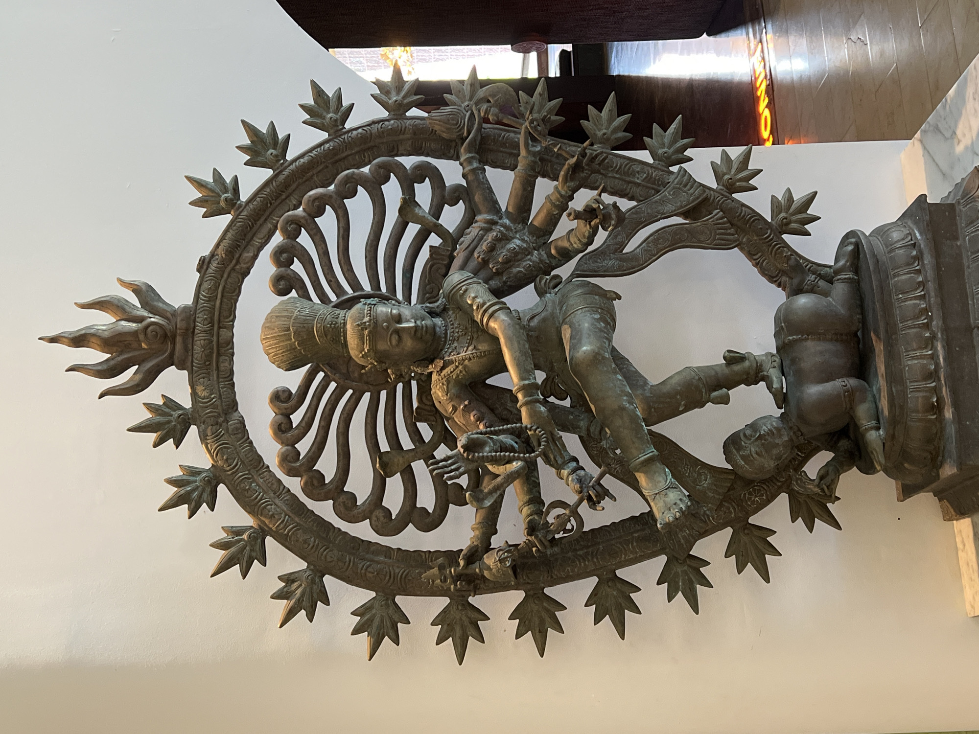 The image shows a bronze statue of Shiva Nataraja, the dancing form of the Hindu god Shiva. It is mounted on a pedestal. Shiva is depicted with multiple arms, dancing within a ring of flames or light. A demon figure lies beneath his foot. The statue appears to be indoors. The photograph was taken in Petulu, Indonesia.