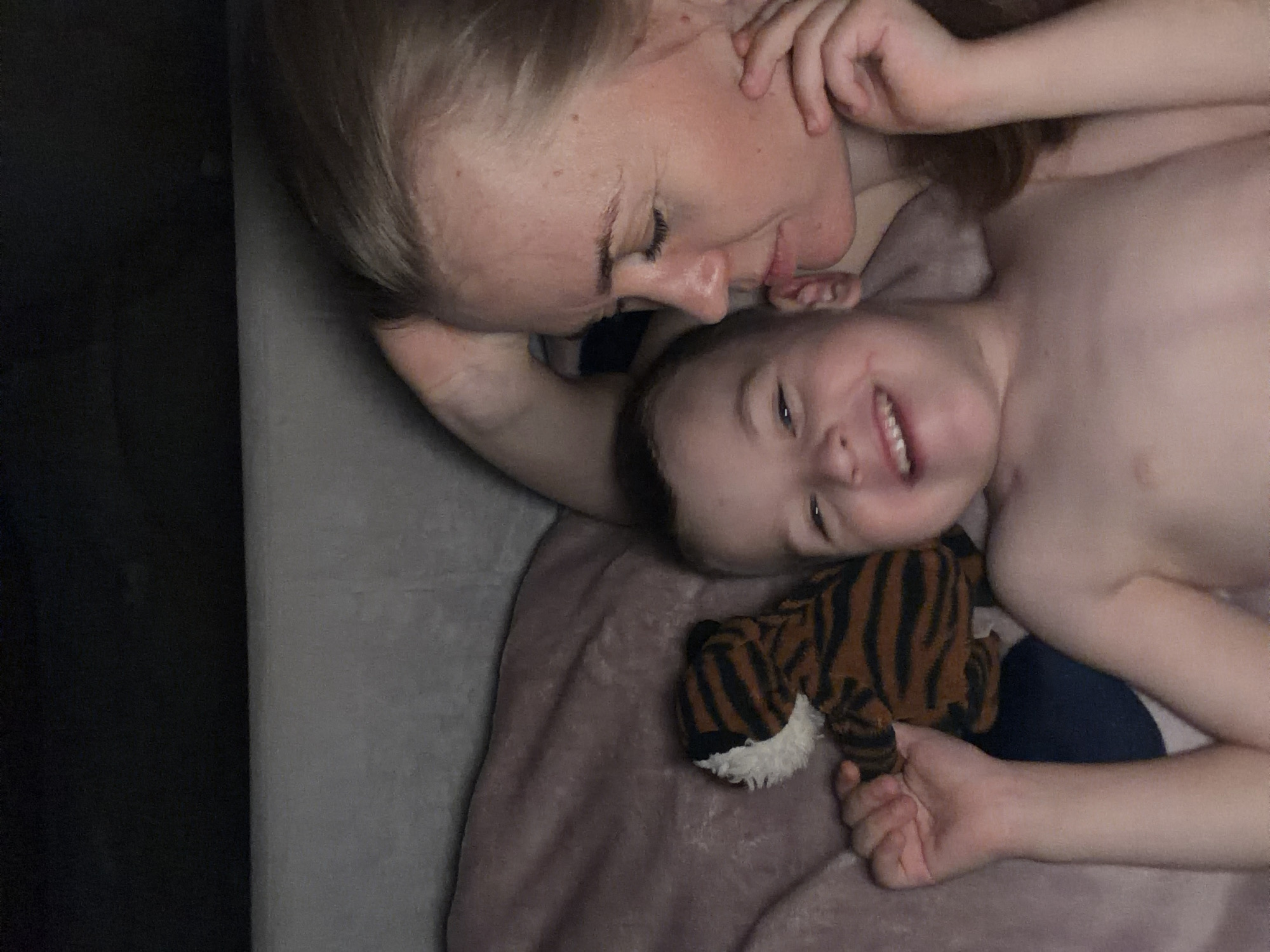 The image shows a mother and her young child lying down, seemingly in bed, given the presence of a blanket and pillow. The mother, with her hair light brown, is on the left side of the frame, looking down at her child with a gentle, affectionate expression. Her right arm is bent and her hand is near her face, perhaps resting or touching her cheek. The child, who appears to be a boy, is on the right, smiling broadly with his mouth open, showing teeth. He is shirtless and has light brown hair. A small, stuffed tiger toy with orange and black stripes lies between the child and the mother's torso. The setting appears to be indoors, in a dimly lit room, suggesting it could be nighttime or early morning. The overall mood conveyed is one of warmth, love, and closeness between mother and child. There is no visible text in the image. The location context provided is Khabarovsk, Russia, but there are no specific visual cues within the image to confirm this location.