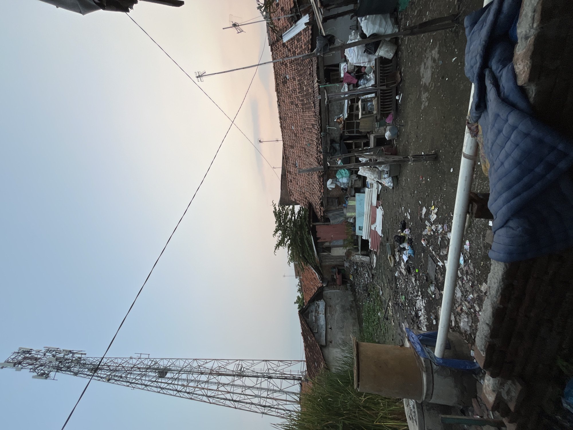 This image depicts an outdoor scene in Pekalongan, Indonesia, likely during the late afternoon or early evening. The foreground is dominated by a pile of dark blue quilted material, possibly a jacket or blanket, resting on a low brick structure. A white pipe runs horizontally across the mid-ground, partially obscured by the brickwork.
Beyond this, the scene reveals a yard littered with a significant amount of trash, including plastic bags, bottles, and other debris. Several structures with red tile roofs are visible, suggesting a residential area. One prominent feature is a tall, metal telecommunications tower situated to the left of the frame, rising above the rooftops. In the background, the sky displays a gradient of light blue and soft orange, indicating either sunrise or sunset. The environment appears somewhat disheveled and impoverished, with visible signs of neglect and waste accumulation.