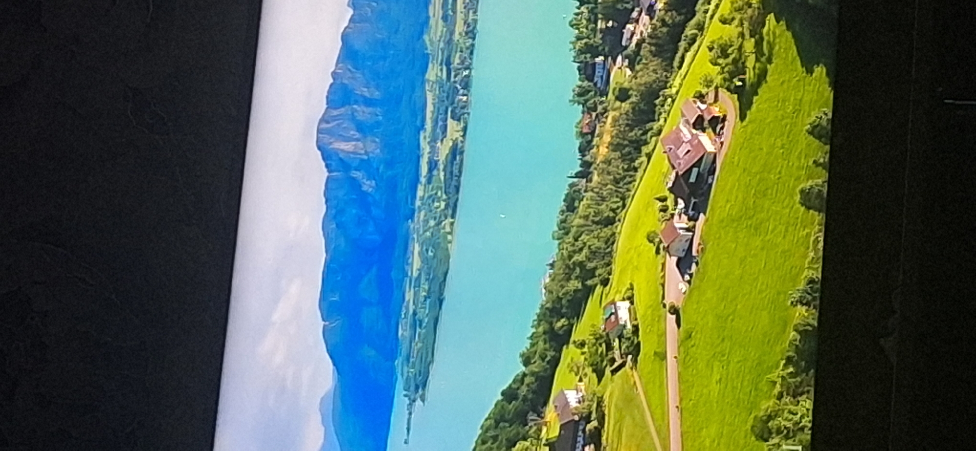 The image depicts a panoramic view of a serene landscape with a large, turquoise lake in the foreground, bordered by lush green hills dotted with houses and trees. In the background, majestic blue mountains rise under a bright, partly cloudy sky. The scene appears to be a rural or semi-rural area, possibly a resort or residential area near a body of water and mountains. There is no indication of the city of Bila Tserkva, Ukraine, in the provided image.