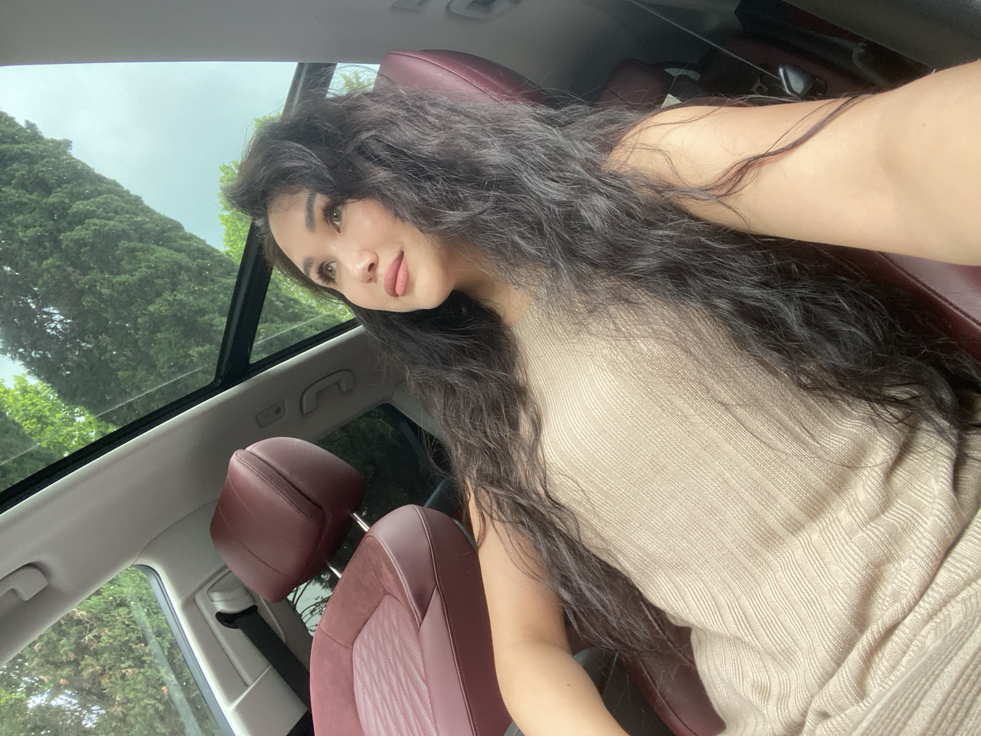 The image is a selfie taken inside a car with a panoramic sunroof. A woman with long, dark, wavy hair is sitting in the driver's seat, angled towards the camera. She is wearing a light-colored, ribbed top. The car has red leather seats. Through the sunroof, lush green trees are visible. Given the presence of lush vegetation and assuming the location provided is accurate, it's likely that the scene is near a park or a forested area in Sochi, Russia. The interior of the car appears clean and well-maintained. The overall impression is one of relaxed leisure.