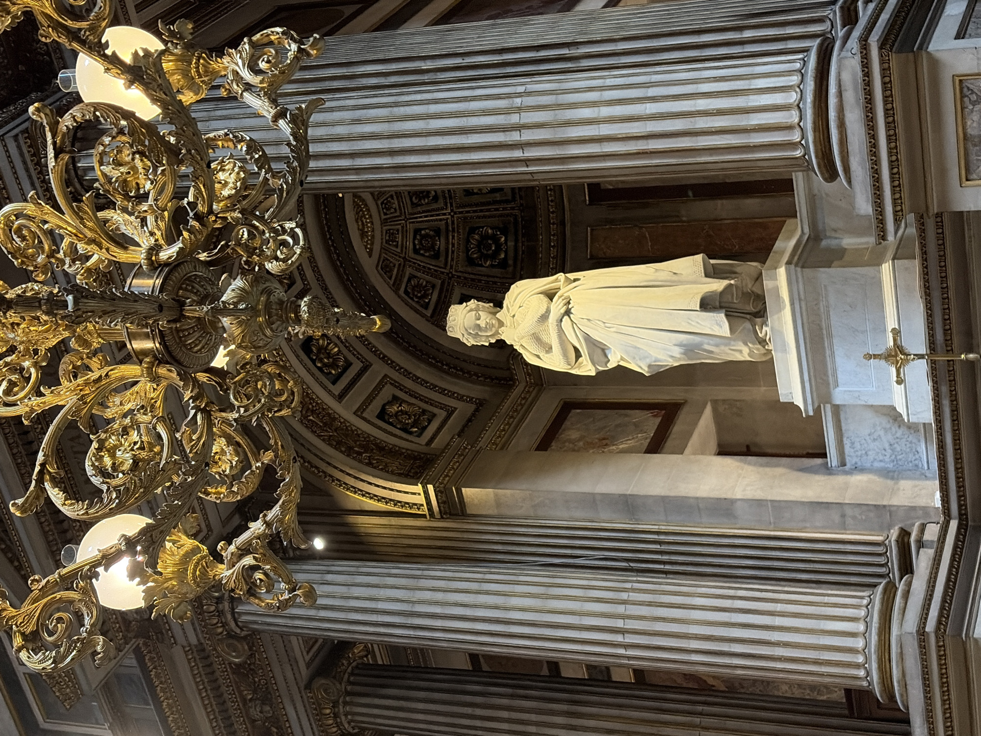 The image depicts a statue of a woman in white marble, dressed in a long robe and wearing a crown, situated within an opulent interior. The statue stands on a pedestal in front of a domed ceiling adorned with intricate gold patterns. The scene is framed by grand, fluted columns with gold accents and a large, ornate golden chandelier hangs overhead, illuminated by glowing lights. A small golden cross is visible at the base of the pedestal. The architectural style suggests a place of religious or historical significance. This statue is likely located in the Sacré-Cœur Basilica in Paris, France, a prominent landmark known for its stunning architecture and religious art.