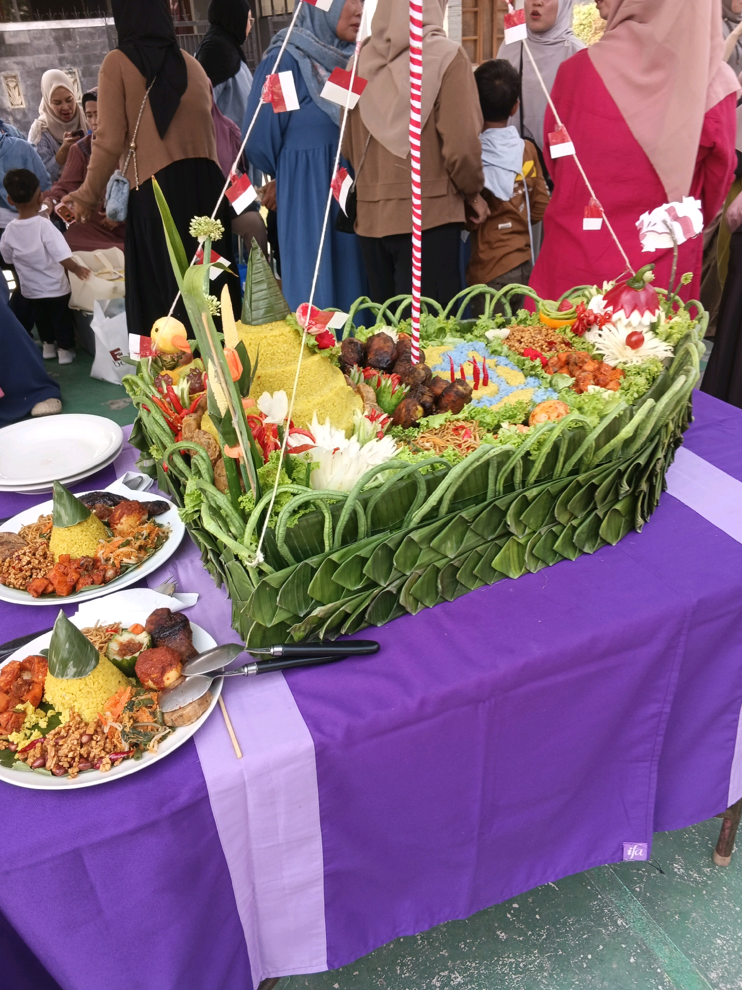 This image depicts a festive outdoor gathering with a prominent food display, likely a celebration or event in Margahayu, Indonesia. In the foreground, two plates of Nasi Tumpeng, a traditional Indonesian cone-shaped rice dish, are laid out on a purple tablecloth. Each plate contains yellow rice, accompanied by various side dishes such as fried chicken, tempeh, vegetables, and noodles.
Behind the plates, a large, elaborately decorated Nasi Tumpeng centerpiece is the focal point. It is shaped like a boat or raft and adorned with fresh green leaves, vegetables, and red chili peppers. Small Indonesian flags are strategically placed around the centerpiece, suggesting a national or patriotic theme.
Several individuals are visible in the background, dressed in traditional attire. Women are wearing hijabs and modest clothing, with one in a bright pink hijab and a matching outfit, and another in a brown hijab and a brown top. Men and children are also present, contributing to the communal atmosphere. The environment appears to be an outdoor setting, possibly a courtyard or street, with a green floor and a wall in the far background. The overall scene conveys a sense of community and cultural celebration, with food playing a central role.