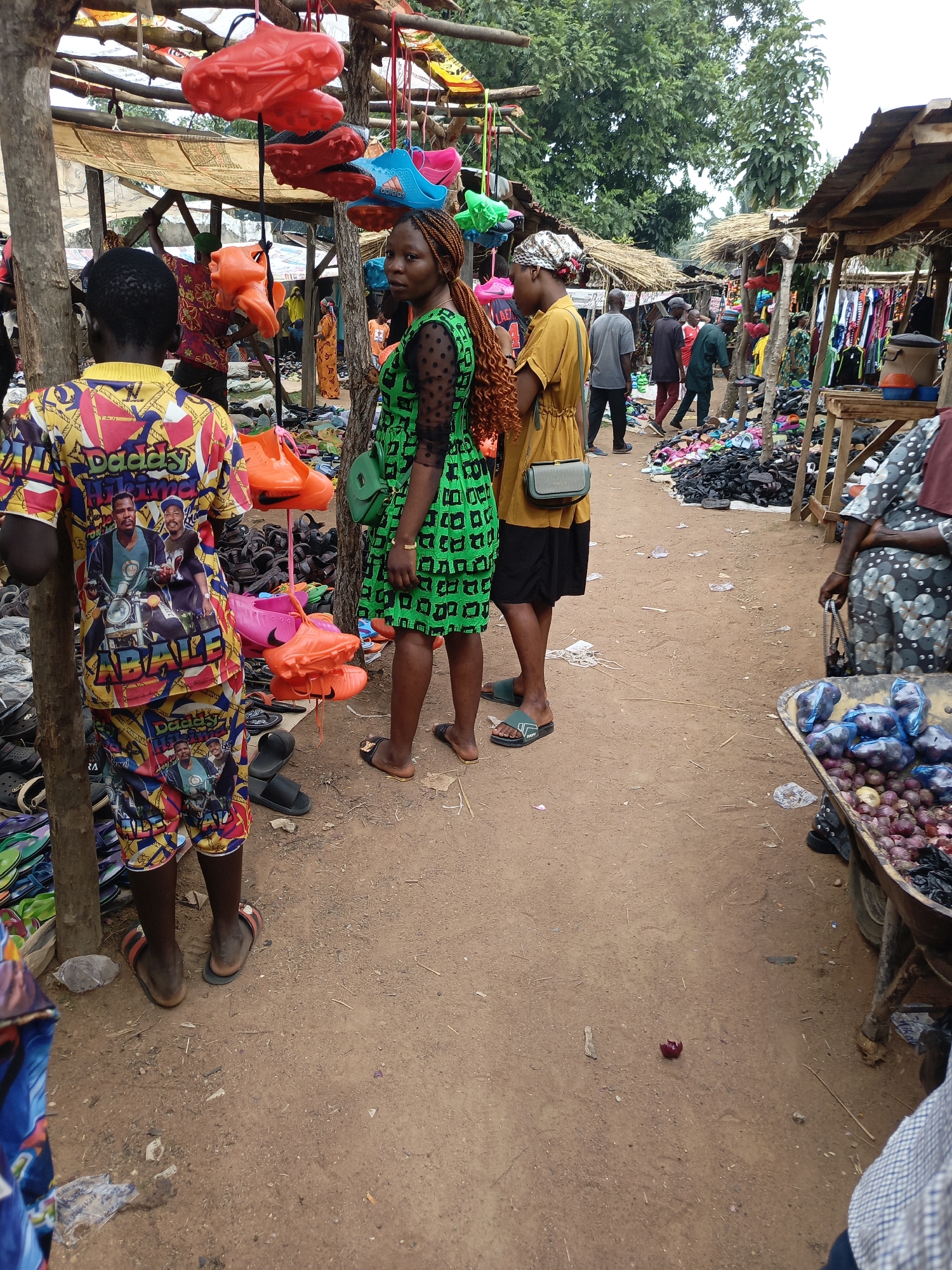 The image portrays a lively open-air market scene in Andaha, Nigeria, during daytime. The ground is unpaved, consisting of light brown dirt, and the market features numerous makeshift stalls constructed with wooden poles and covered with a mix of fabric and corrugated iron sheets.
In the immediate foreground on the left, the back of a person wearing a vibrantly patterned, multi-colored shirt and matching shorts is visible. The clothing features abstract designs and portraits of two men, with the prominent text "DADDY ABALE" printed across it. This individual stands next to a wooden post from which several pairs of bright red, orange, and pink shoes, resembling clogs or slippers, are hanging for sale.
Slightly to the right, two women stand on the dirt path. One woman, with long, reddish-brown braided hair, wears a green dress with a dark square pattern and sheer, polka-dotted sleeves. She looks directly towards the viewer and carries a small green cross-body bag. Beside her, a second woman, facing away, wears a mustard-yellow top, a black skirt, and a light-colored headscarf, carrying a grey cross-body bag.
The market path recedes into the background, lined with more stalls. To the right, a large pile of various shoes, predominantly dark-colored, is spread on the ground. Further back, clothing is displayed on racks. On the far right, a robust wheelbarrow or cart is filled with red and white onions, some bundled in blue plastic bags, indicating a produce section. A single piece of red onion peel lies on the ground.
Numerous other people are visible in the midground and background, walking, browsing, and engaging in market activities. Green trees are visible above the market structures, suggesting an outdoor, possibly rural setting. On one of the blue hanging shoes, the brand name "Adidas" is faintly discernible. The scene captures the essence of a bustling local market.
