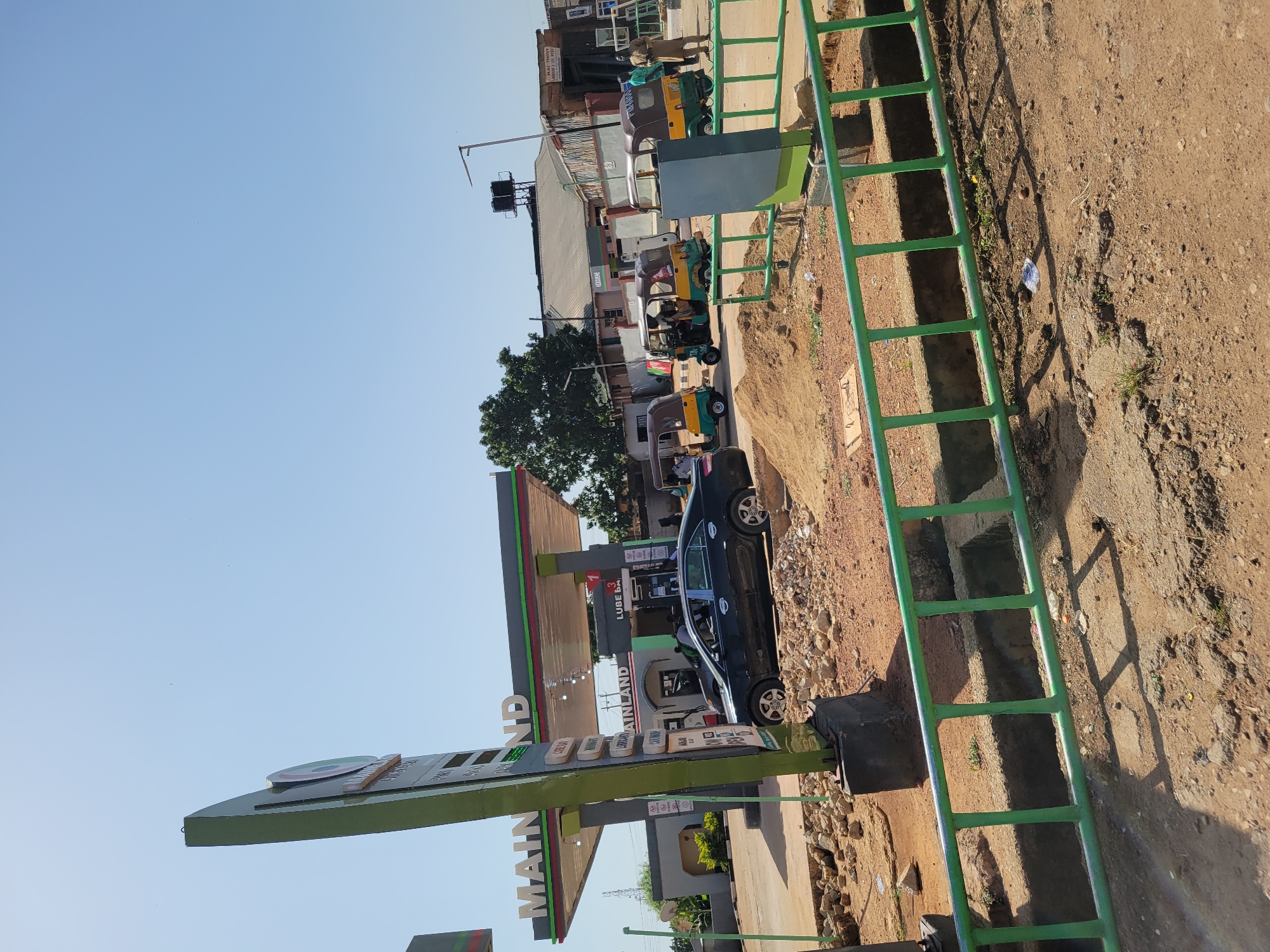 A street scene in Unknown, Unknown, displays a green Main Land gas station with a black car parked at a pump. Several three-wheeled vehicles are visible down the street. A green painted metal ladder-like structure is positioned on the right side of the frame on a dirt shoulder. The sky above is clear and blue.