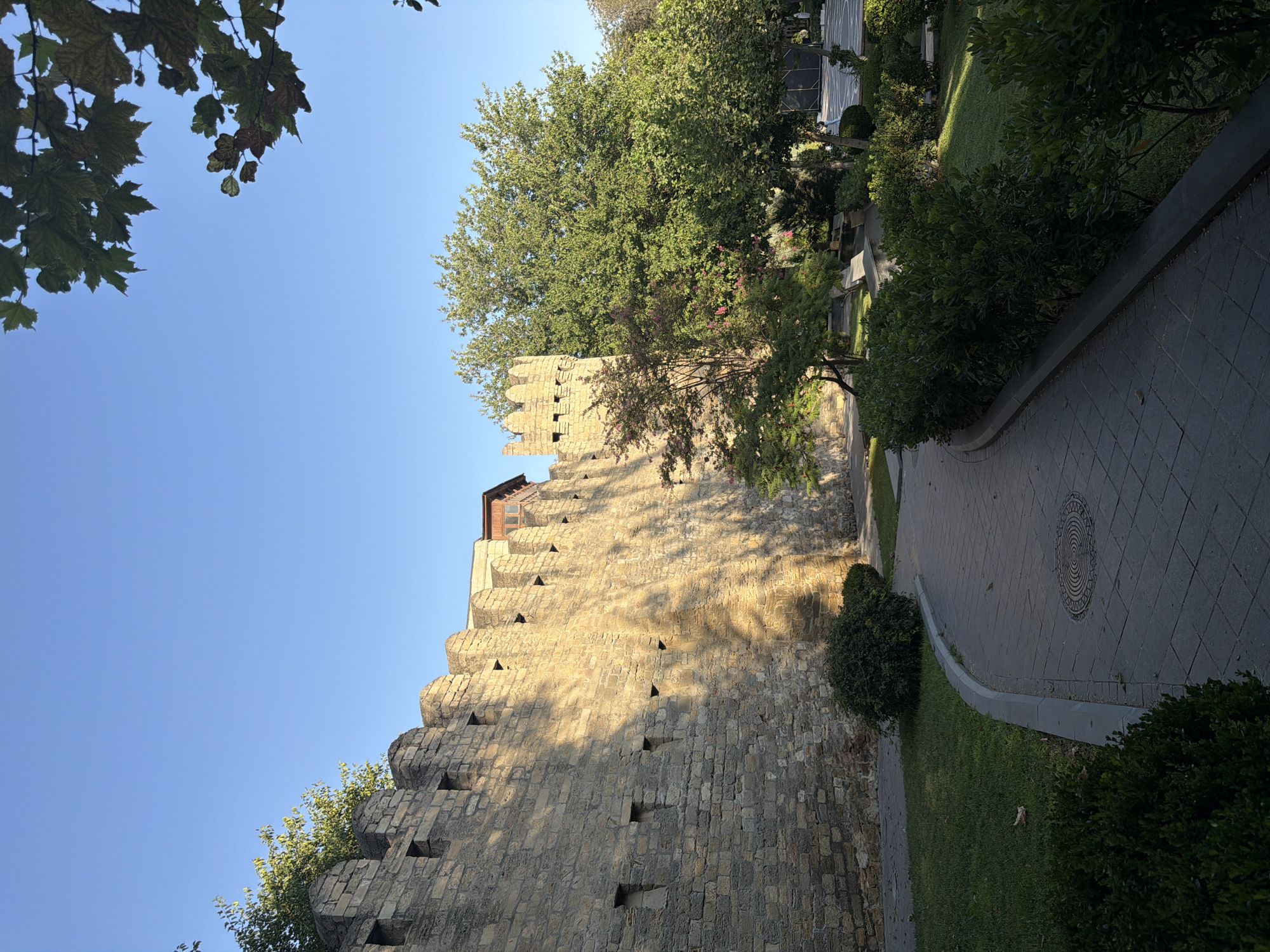 The image depicts the Maiden Tower, a historic fortress located in the Old City of Baku, Azerbaijan. The tower is a cylindrical stone structure with crenellated battlements at the top, and the image shows a section of its imposing stone wall extending to the left. The wall is constructed from light-colored, rough-hewn stones, with rectangular openings visible at intervals. Sunlight casts a dappled shadow pattern across the wall, suggesting it is mid-morning or late afternoon.
In the foreground, a paved walkway with a textured surface curves to the right. The walkway is bordered by neatly trimmed green bushes and a strip of manicured lawn. Beyond the walkway and bushes, there is a park-like area with more trees and shrubbery, some of which are lush and green, while others show signs of autumn coloration. The sky above is a clear, bright blue. The overall impression is of a well-maintained historical site with a park setting. There are no people visible in the image.