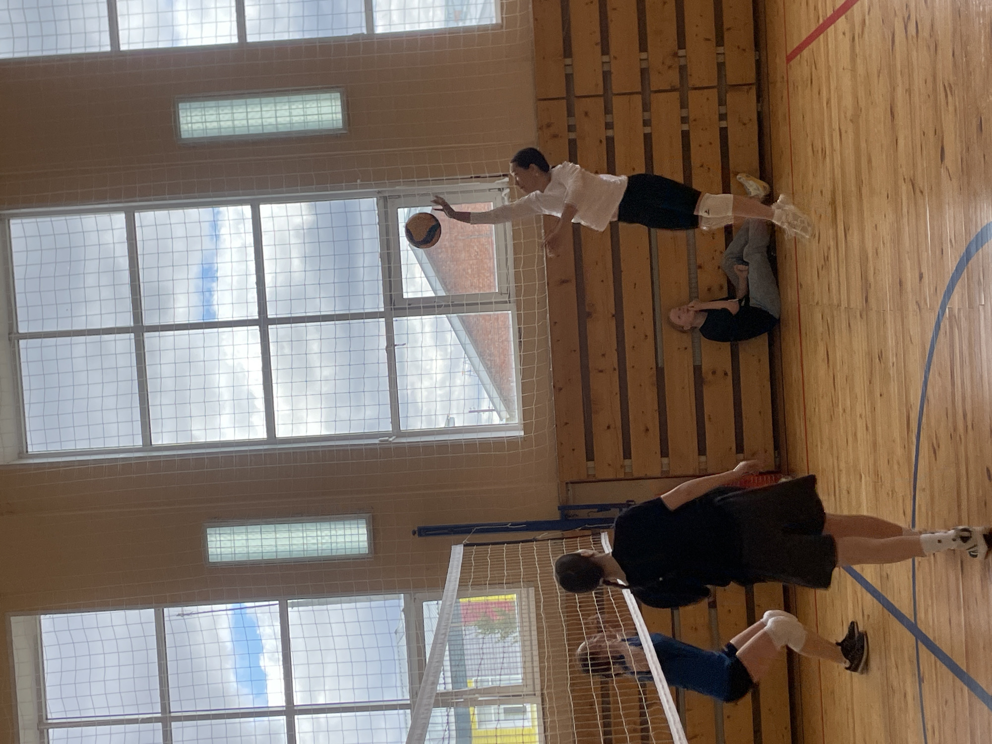 A gymnasium in Подшивалово, Russia, features a volleyball game. Two players are near the net on one side, one in mid-jump, the other crouching. On the opposite side, a player leaps towards a volleyball, while another lies on the floor. Large windows line the wall, revealing a cloudy sky. The gymnasium has a wooden floor with blue lines, a volleyball net, and light fixtures.