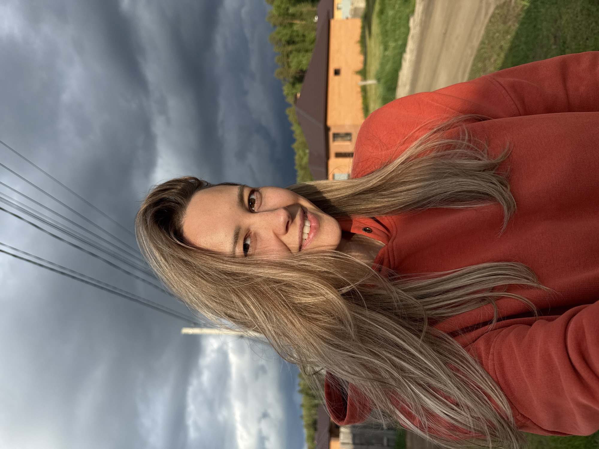 A fair-skinned woman with long, light-colored hair is smiling at the camera. She's wearing an orange-red sweater. Behind her is a gray, cloudy sky and a view of a brown brick house and green foliage, suggesting an outdoor setting. The presence of power lines in the background implies a residential area. Based on the location provided, the scene is likely in Chaikovsky, Russia, but precise coordinates would be needed to confirm the exact street or neighborhood. There are also trees visible at the top of the image. A road is seen near the brown brick building.
