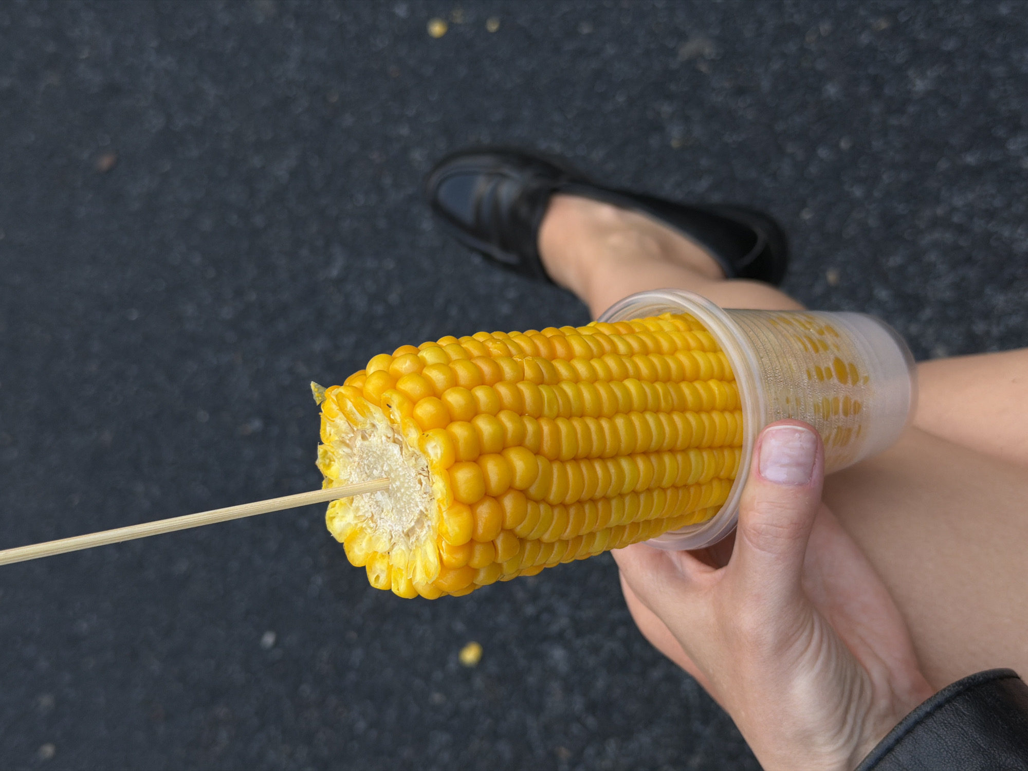 An aerial shot shows a woman holding a cup of corn in her left hand while sitting outside. The corn is on a stick and the cup contains some liquid, possibly butter or water. The woman's legs are visible, and she is wearing black shoes. The background is asphalt. The location of the image is unknown and the city of Saratov, Russia is not discernible in this image.