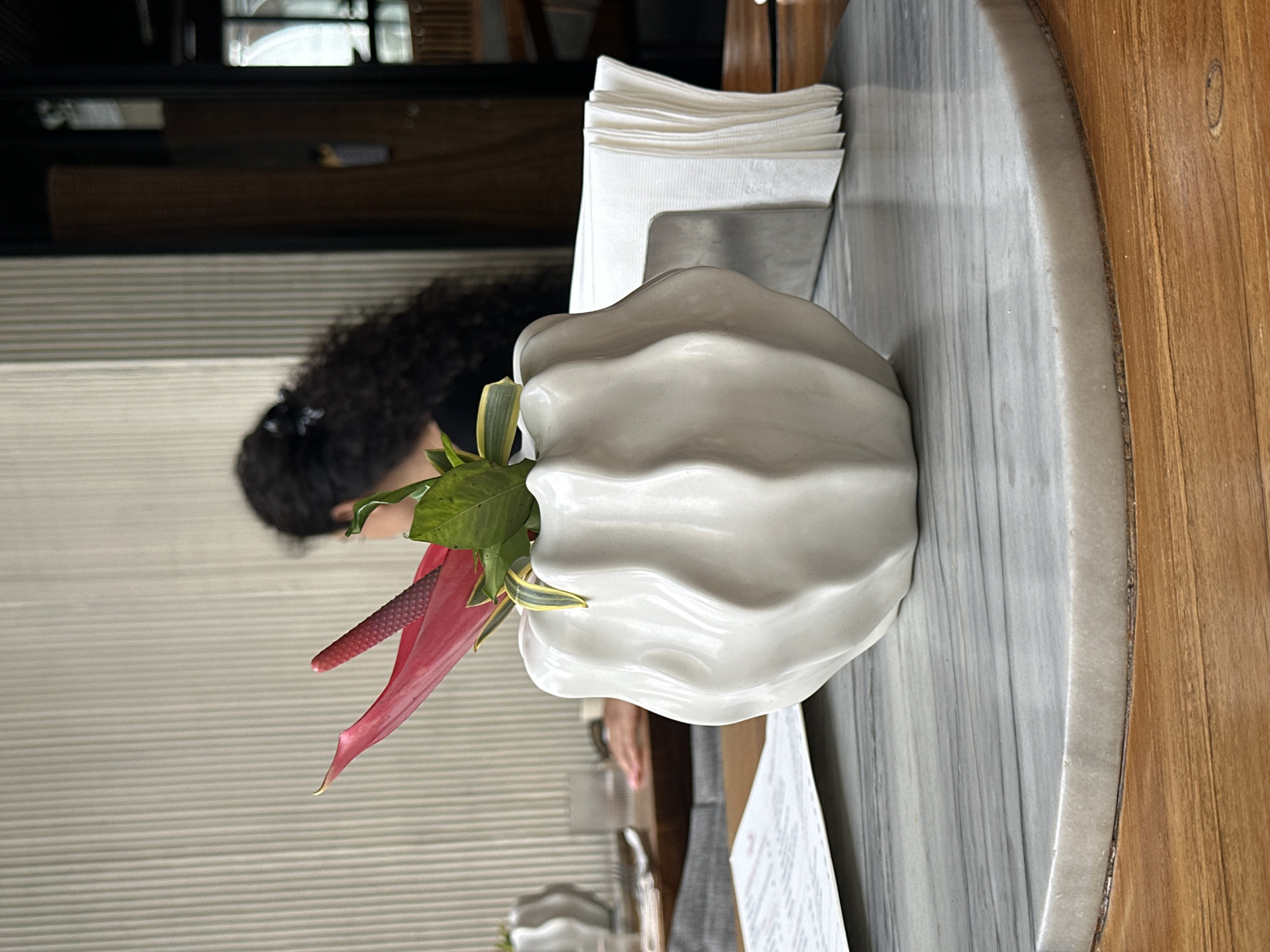 In North Kuta, Indonesia, a table setting features a white, wavy vase with a vibrant pink anthurium flower and green leaves. To the right of the vase is a metallic napkin holder filled with white napkins. In the background, a woman with long, curly dark hair is visible from behind, wearing a black top. The table is round and appears to be made of marble on top of a wooden base. A menu is placed in the foreground, slightly out of focus. The wall behind the setting has a ribbed texture.