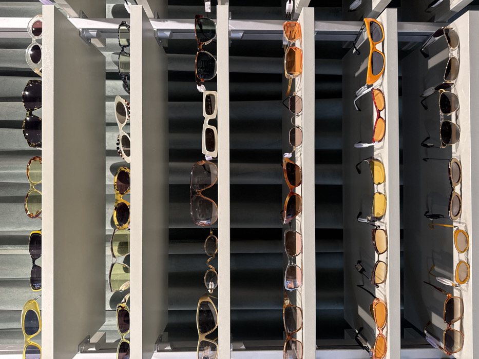 This image depicts an indoor retail display of numerous sunglasses arranged on multiple white shelves against a background of vertical, pleated, dark green fabric curtains. The scene is located in Paris, France.
Approximately 60-70 pairs of sunglasses are visible, neatly organized in rows on at least eight distinct shelves. The sunglasses exhibit a wide variety of styles, frame shapes (including aviator, cat-eye, oval, rectangular, and oversized designs), frame colors (ranging from solid black, white, and yellow to tortoiseshell and transparent hues), and lens tints (including dark, mirrored, and gradient shades of brown, grey, green, and orange). Each pair of sunglasses is positioned individually on the shelves, with some frames featuring price tags or branding labels attached to their temples or lenses. The shelves themselves are thin, flat, and appear to be made of a light-colored material, likely white or off-white, supported by subtle vertical structures on the right side of the frame. No individuals, explicit actions, or direct interactions are visible within the frame. The lighting appears even, suggesting an indoor, well-lit environment.