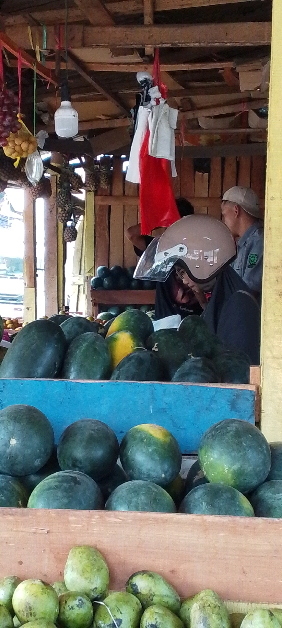 A street market stall in Cilaku, Indonesia, displays a variety of fruits, primarily watermelons and some green mangoes or papayas. In the background, a man wearing a light-colored cap and a dark shirt with a green safety emblem is visible. To his left, another person, wearing a helmet with a clear visor, is seated. The stall itself is constructed from wood, with a corrugated roof. Red and white fabric, possibly a flag, hangs from the ceiling, along with a bare light bulb. Bunches of red grapes and other small, round fruits are also present in the upper left portion of the image. The overall impression is of a vibrant, local marketplace.
