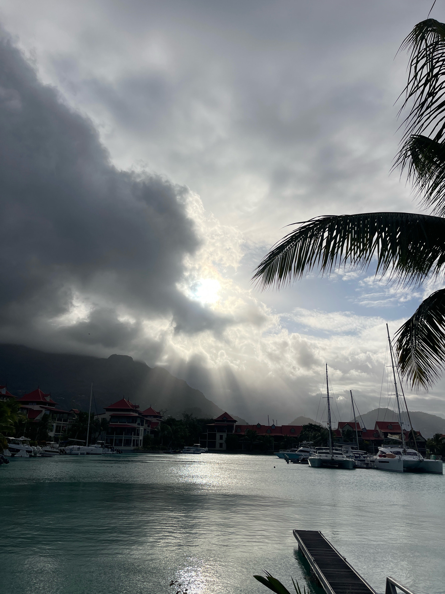The image shows a harbor in Providence, Seychelles, with several sailboats and catamarans docked. The sun is setting, casting rays of light through the clouds and illuminating the water. Palm trees frame the scene, and mountains can be seen in the background. The architecture of the buildings suggests a resort or hotel complex. The water is calm, reflecting the light and creating a shimmering effect. There are no people visible in the image. The overall atmosphere is serene and tranquil.
