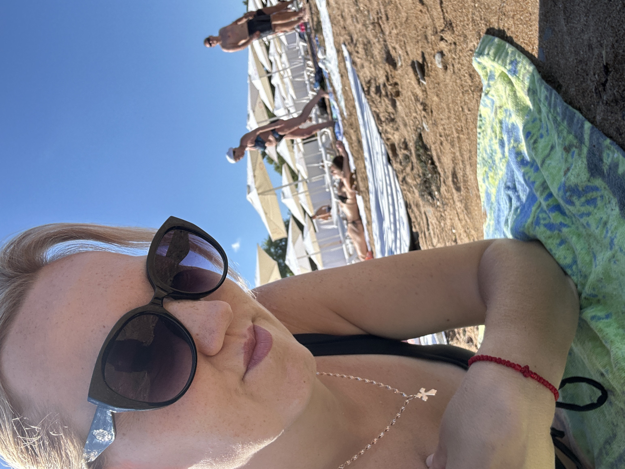 The image shows a woman wearing sunglasses and a black bikini top lying on a towel on a sandy beach. She is wearing a necklace with a cross pendant and a red beaded bracelet. In the background, several beach umbrellas and loungers are visible, as well as other people in swimsuits. The sky is clear and blue. The scene appears to be a resort beach, likely in the city of Gelendzhik, Russia, given the context provided.