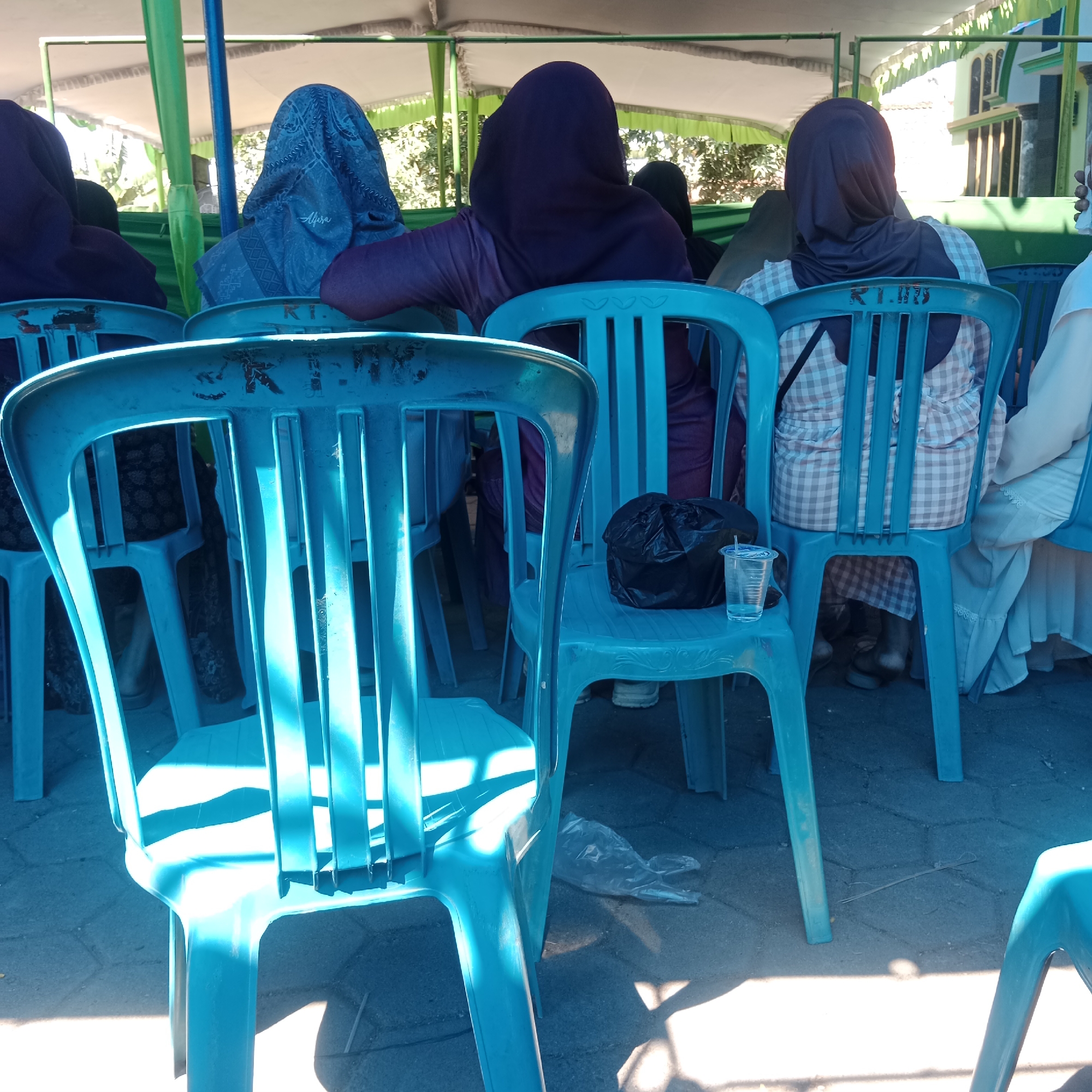 In the foreground, a bright blue plastic chair is partially visible, casting a strong shadow on the grey paved ground. Beyond it, a row of similar blue plastic chairs stretches into the background. Several individuals, primarily women, are seated on these chairs, facing away from the camera. They are all wearing headscarves, with some in shades of purple and blue. One woman in the mid-ground wears a blue patterned headscarf and a checkered top. Another, further back, has a dark purple headscarf and top. A black plastic bag rests on the seat of a chair in the mid-ground, next to a clear plastic cup with a straw. In the background, green and white tarpaulin structures are visible, supported by poles. Beyond the tarpaulin, trees and a building with arched windows can be seen, suggesting an outdoor gathering or event. The scene appears to be set in Tegowanu Kulon, Indonesia, likely a community event or religious gathering due to the attire of the attendees.
