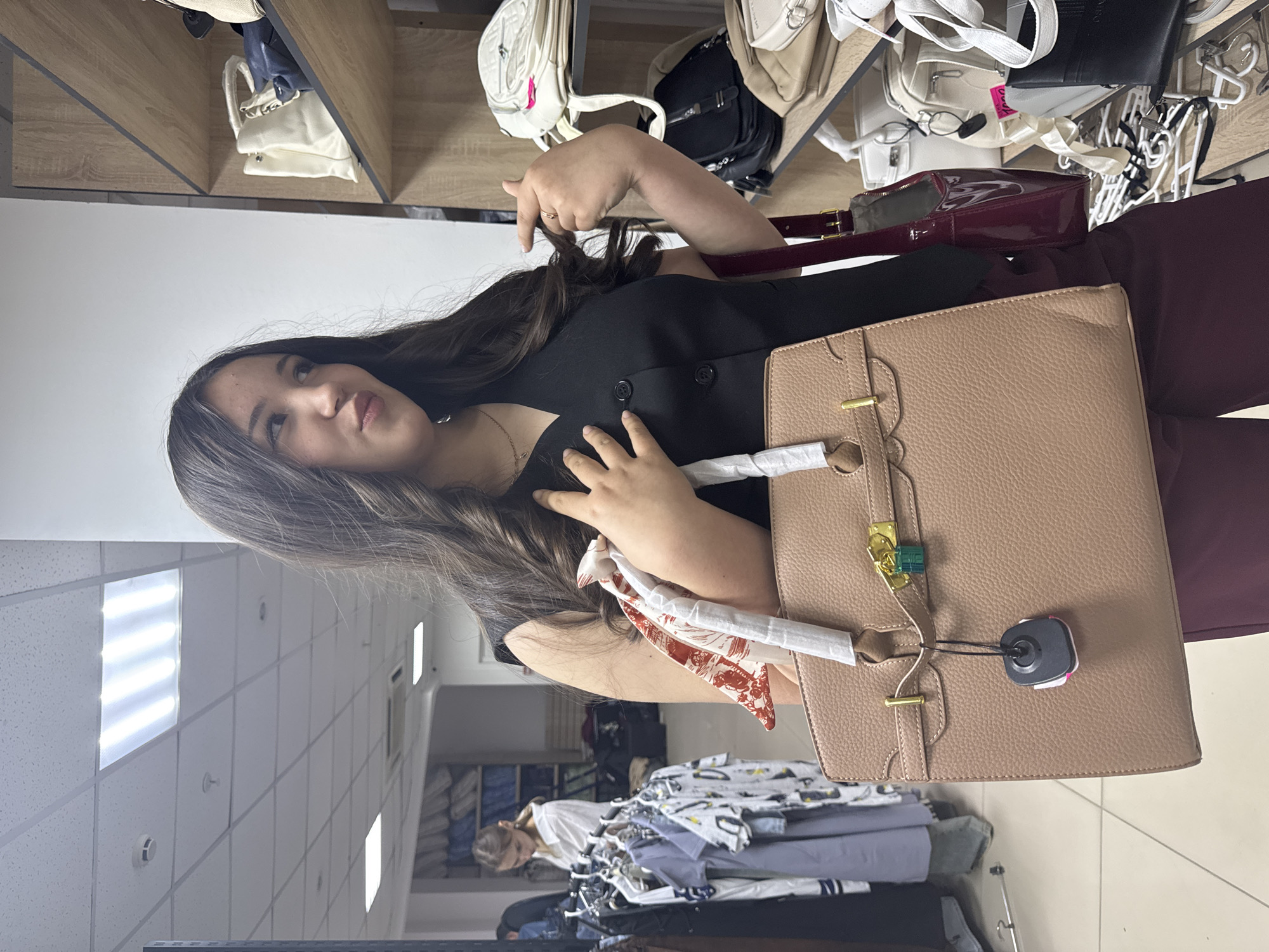 A young woman is standing in a store, looking to her right with a playful expression. She is wearing a black sleeveless vest over a dark top and dark maroon pants. She is holding a large, light brown textured handbag with a gold clasp and a green padlock. A red and white patterned scarf is tied to the handbag's handle. She is also wearing a necklace with a small pendant. Behind her, to the left, a rack of clothing is visible, with another woman in a white shirt browsing the items. To the right, shelves are stocked with various handbags. The scene appears to be indoors, likely a retail environment, given the presence of clothing racks and merchandise shelves. The image quality suggests it was taken with a mobile phone. No specific details related to the city of Perm, Russia, are discernible from the image itself.