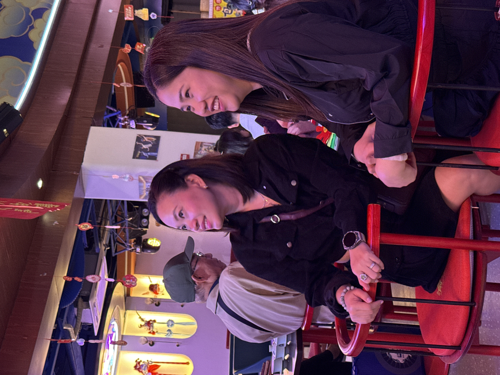 A group of people are on a ride at an amusement park or carnival in Yuzhong District, China. Two young women are in the foreground of the shot on the ride, both wearing dark clothing. One woman is wearing a jacket and smiling, while the other woman is wearing a button-up shirt. In the background, there is a man with a cap and a beige and black jacket. The ride is red and is located in an area with decorations.