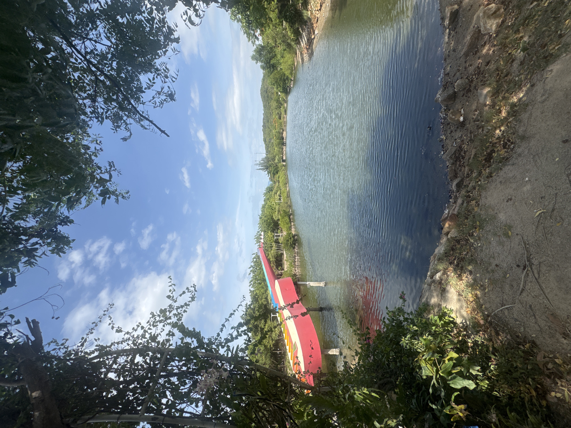In Hoa Thang, Vietnam, a bright red and yellow waterslide juts out over a calm, blue body of water. The waterslide, supported by several sturdy white pillars, appears to be shaped like a guitar. The surrounding environment is lush and green, with trees and foliage framing the scene. The sky above is a clear blue, dotted with fluffy white clouds, suggesting a pleasant day. The water's surface is gently rippled, reflecting the sky and surrounding greenery. The banks of the water are lined with rocks and dry leaves, with more vegetation visible in the background, including distant hills. The overall impression is one of a tranquil, natural setting enhanced by a playful, man-made structure.