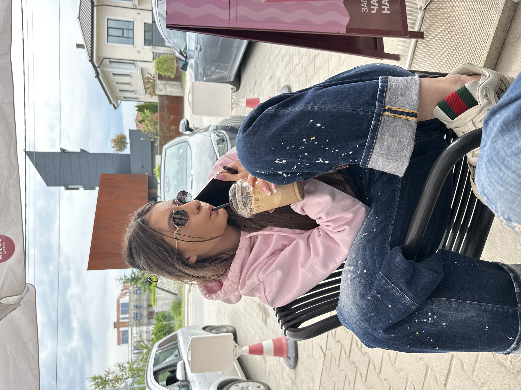 A woman in sunglasses and a pink hoodie is sitting on a black metal bench at a table, drinking from a plastic cup with a straw. She is wearing jeans with paint splatter details and white sneakers with red and green stripes. She is holding a black smartphone in her left hand. The location appears to be an outdoor cafe in Plovdiv, Bulgaria, as evidenced by the architectural style of the buildings in the background. Cars are parked on the street behind her. A traffic cone is on the ground next to a white signpost, and another table with a pink sign is visible in the background.