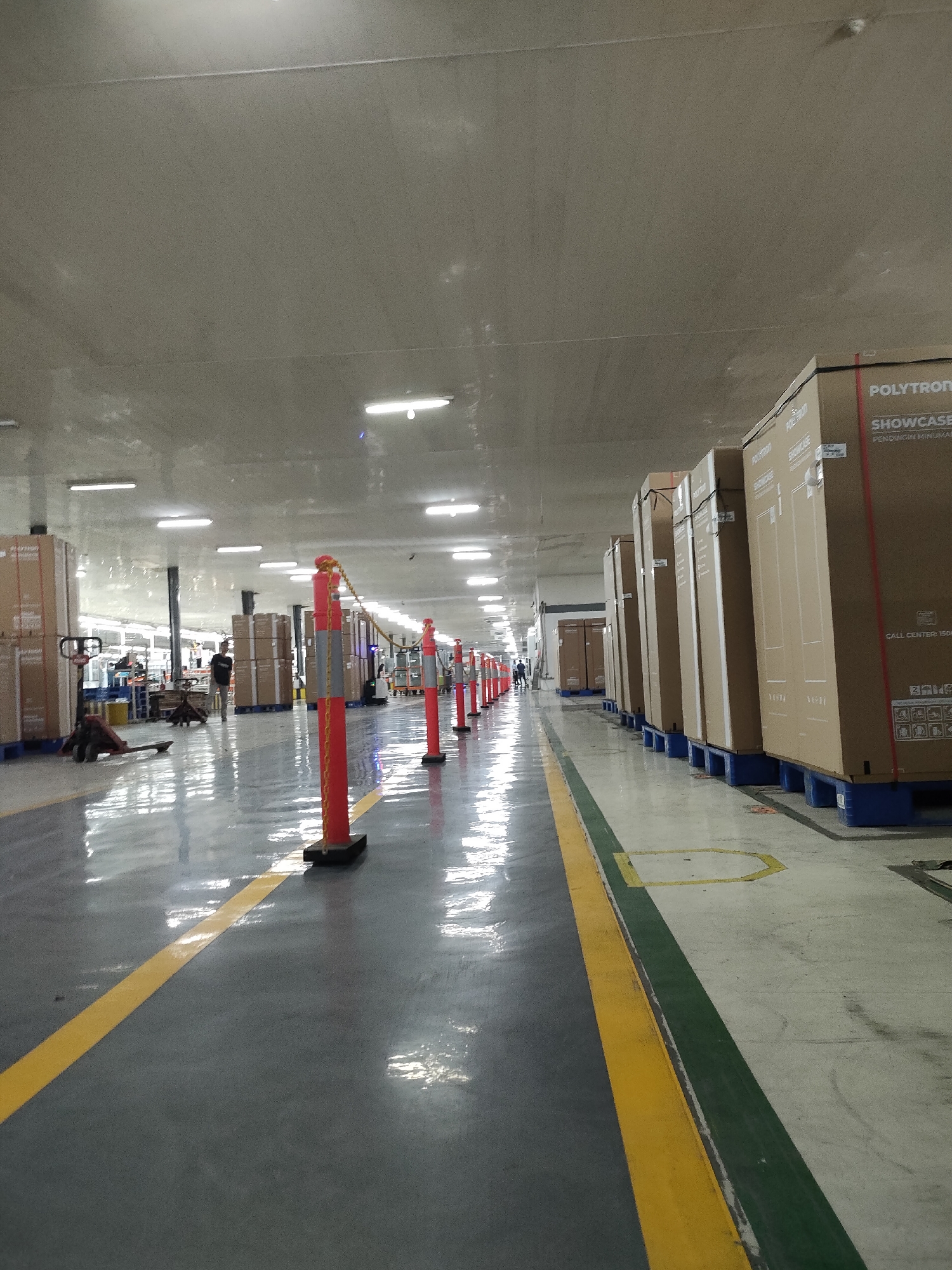 The image depicts the interior of a warehouse located in Sayung, Indonesia. The warehouse features a high ceiling with fluorescent lighting. The floor is a polished concrete surface with yellow and green painted lines indicating pathways. Rows of large, cardboard boxes, many labeled "POLYTRON SHOWCASE," are stacked on blue pallets throughout the warehouse. In the foreground, red and grey bollards with yellow chains mark a boundary. A person wearing dark clothing is visible in the middle distance, walking away from the camera. In the background, other workers and equipment, including a pallet jack, can be seen. The overall impression is that of an organized, active industrial or distribution facility.