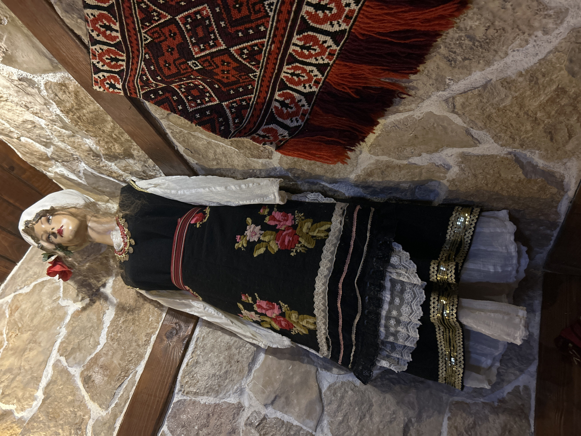 The image shows a traditionally dressed doll displayed against a stone wall, with a woven rug hanging above it. The doll has fair skin, red lipstick, and is wearing a white headscarf, a red rose, and a traditional black dress with floral embroidery and lace trim. The dress also features gold sequins at the bottom. A red and black patterned rug with fringe hangs behind the doll, partially covering the stone wall. The scene is likely inside a building with a stone interior. The location is specified as Mirovyane, Bulgaria, based on the prompt.