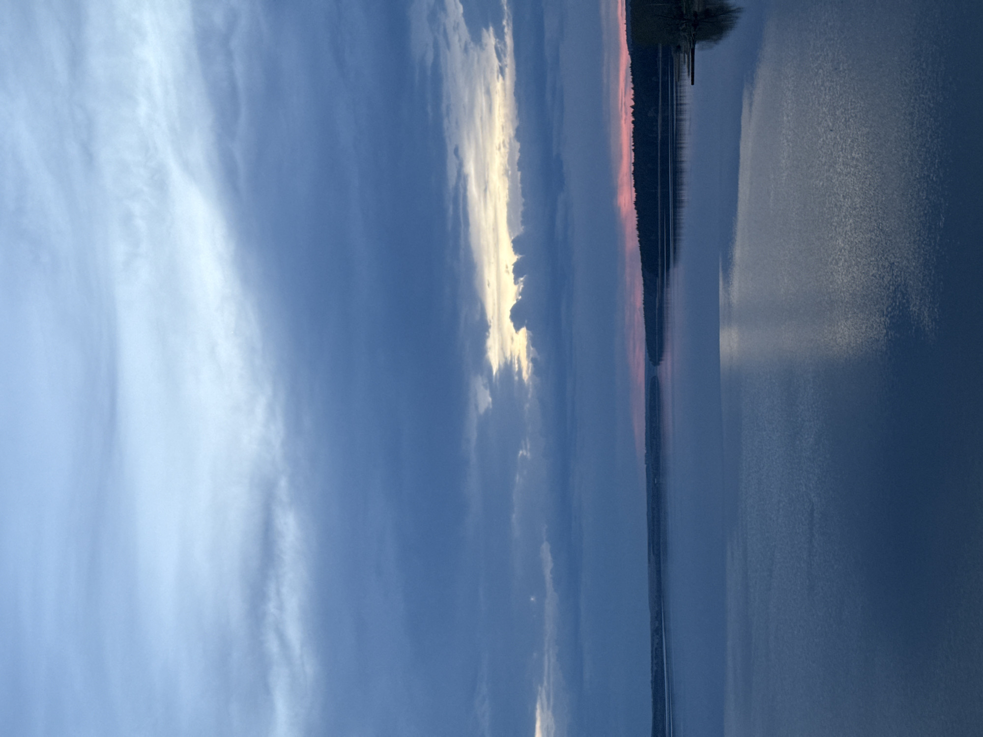 The image depicts a body of water, possibly a lake or river, with the sky above. The sky shows a variety of cloud formations, ranging from light, wispy cirrus clouds to darker, more dense cumulonimbus clouds. A bright, almost golden light shines through a gap in the clouds. A thin strip of land, possibly a forest, is visible in the distance, with a pink hue suggesting either a sunrise or sunset. The water reflects the colors of the sky. The image's orientation is vertical. The location of the image is unclear, and it's difficult to determine if it is indeed from Votkinsk, Russia. There are no people visible in the image.
