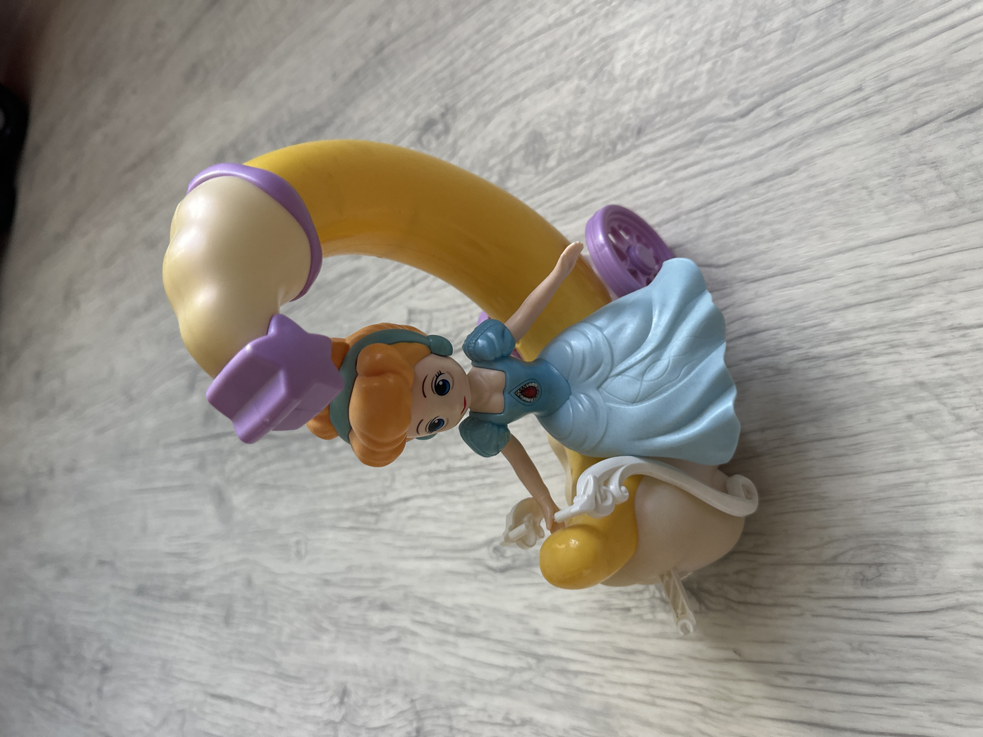 A plastic figurine of a princess, resembling Cinderella, sits on a yellow and purple carriage. The princess wears a blue gown and has orange hair. The carriage has a curved yellow armrest, a purple wheel, and a tan base with white string attached. The figurine is positioned on a light gray wooden floor with visible grain patterns. The scene is devoid of any urban or specific geographical context related to the city of Мариинский, Russia.