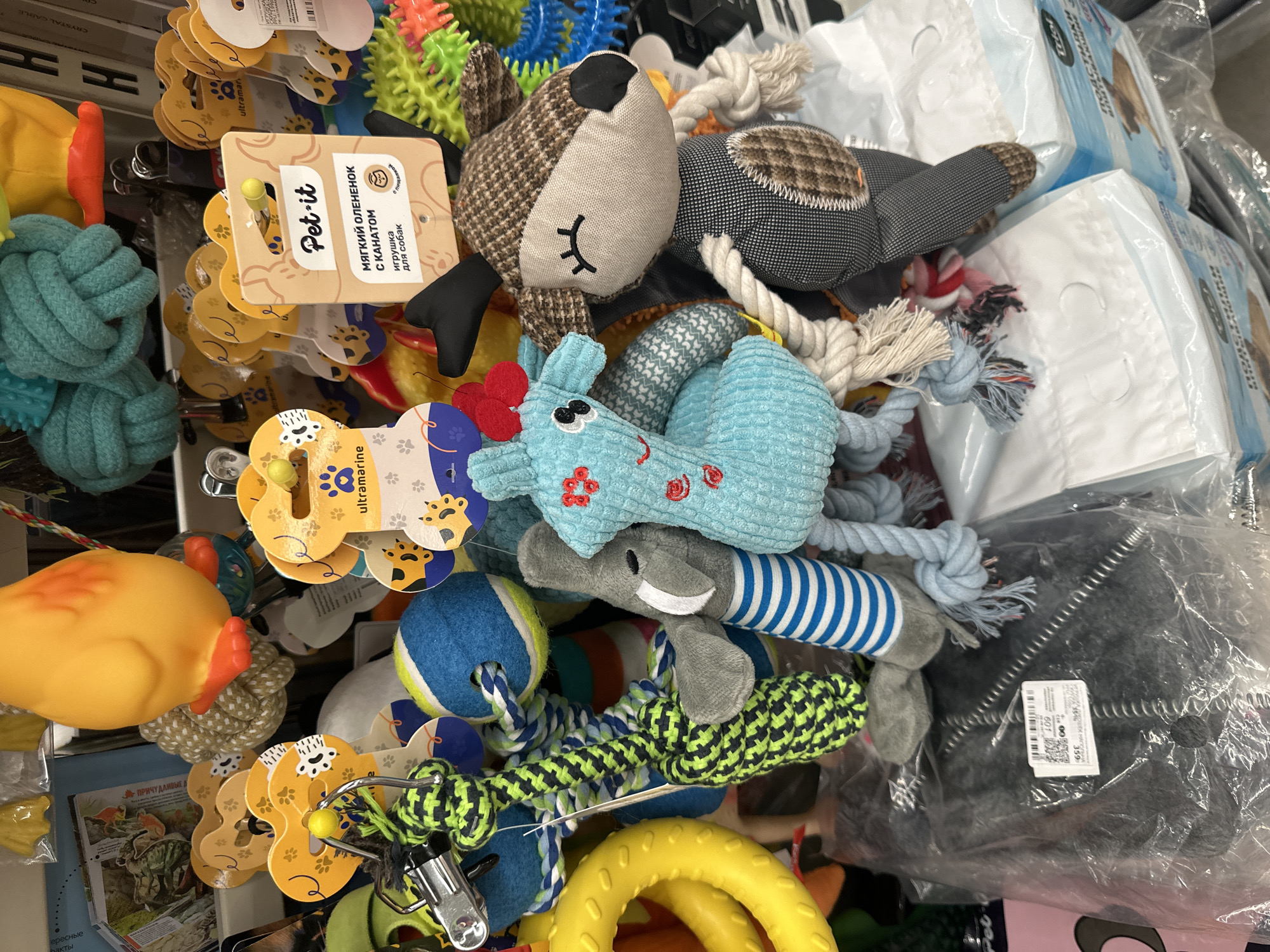 The image displays a close-up, eye-level view of a retail display of dog toys in an unknown location. The foreground is dominated by various plush and rope dog toys, including a brown and white plaid deer with embroidered closed eyes, a blue plush giraffe-like toy with red accents, and a gray plush elephant with blue and white striped legs. Several rope toys with knotted sections are also visible, along with a yellow ring toy and a blue and yellow tennis ball. The background features more colorful dog toys, including some spiky plastic ones. Packaging for what appears to be dog diapers or puppy pads is stacked in the lower right corner, with text partially visible in Russian. A price tag with Cyrillic script and a barcode is attached to a plastic-wrapped item in the lower left. The lighting suggests an indoor retail environment.