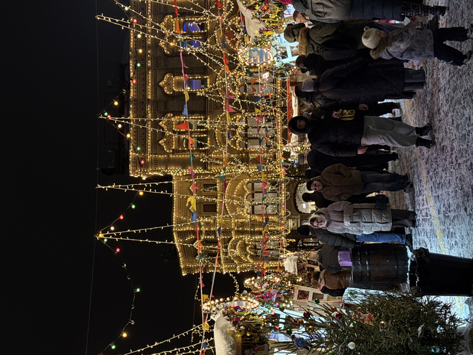 The image displays a nighttime winter market scene situated in Moscow, Russia, directly in front of the GUM (State Department Store). The ground is covered with a layer of snow, indicative of cold temperatures. A crowd of individuals, dressed in heavy winter coats, hats, and scarves, occupies the foreground and midground, moving and standing throughout the market space.
The multi-story GUM building dominates the background, extensively illuminated by warm white string lights that outline its windows, architectural features, and roofline. Colorful triangular bunting flags are strung across the building's facade. Overhead, numerous strings of spherical multicolored lights (red, green, blue, yellow) are suspended, crossing the open market area.
In the lower left foreground, a small, decorated evergreen tree stands next to a black trash receptacle. A wooden barrel is visible near a market stall on the left, which is also adorned with lights and shows accumulated snow on its roof. A young child in a patterned winter coat and hat walks towards the right in the lower right foreground. Adults are observed walking, observing, and mingling within the crowded environment.