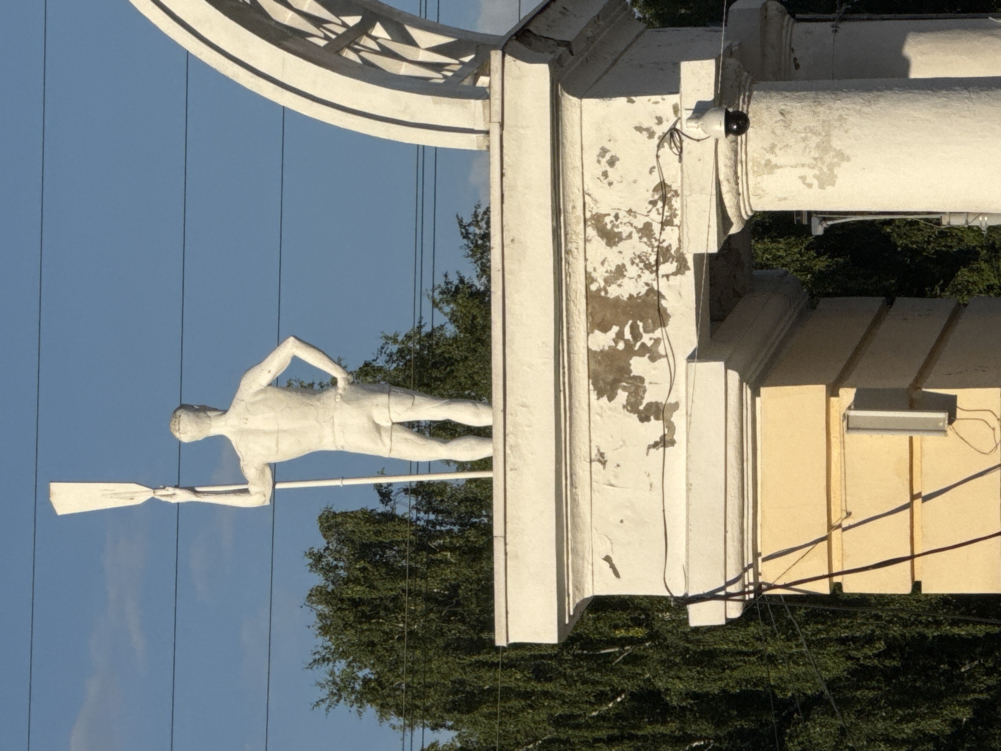 The image shows a white statue of a man holding a paddle, standing on top of a white architectural structure. The statue faces away from the camera, with its right hand raised holding the paddle. The man is wearing shorts. The structure it stands on has a textured, peeling surface. To the right of the statue, a white arched structure with geometric patterns is visible. In the background, there are green trees and a clear blue sky with power lines. A security camera is mounted on one of the columns supporting the structure. This statue is located at the entrance to the Izhevsk Water Stadium, an outdoor sports complex in Izhevsk, Russia, dedicated to water sports and recreation.
