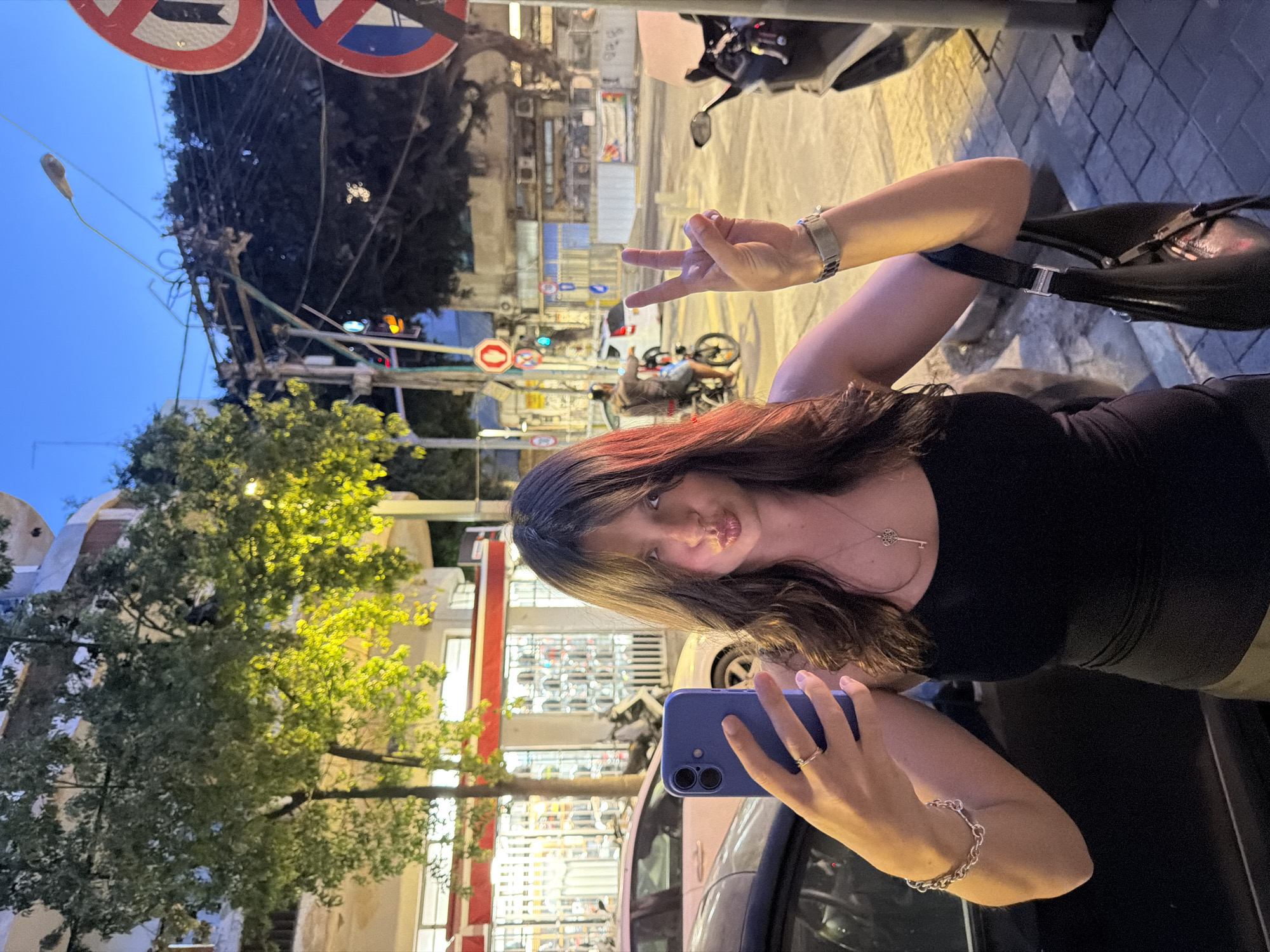 A woman is taking a selfie at night in Tel Aviv, Israel. She is standing next to a dark-colored car and holding a blue smartphone in her right hand. She is wearing a black top and a silver bracelet on her right wrist. She is making a peace sign with her left hand. The background shows a typical Tel Aviv street scene with buildings, streetlights, traffic signs (including a "no entry" sign), and a tree with green foliage. There is a scooter parked on the street in the background. The overall lighting suggests it is dusk or early evening.