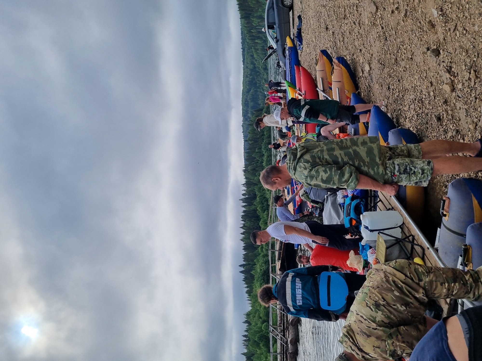 The image depicts a group of people gathered by a body of water, possibly preparing for or concluding a rafting trip. Several inflatable rafts of various colors, including red, blue, and yellow, are present on the shore and in the water. The shore is covered with pebbles. In the background, a dense forest stretches along the opposite bank of the water. The sky is overcast, with some sunlight breaking through the clouds in the upper left corner. A dark-colored car is parked on the right side of the image. People are seen in casual clothing, some of them interacting with the rafts or carrying equipment. One individual is wearing a jacket with "СИБУР" written on the back, a prominent Russian petrochemical company. The scene suggests an outdoor recreational activity taking place in a natural, possibly remote, setting near the city of Vishegorsk, Russia.