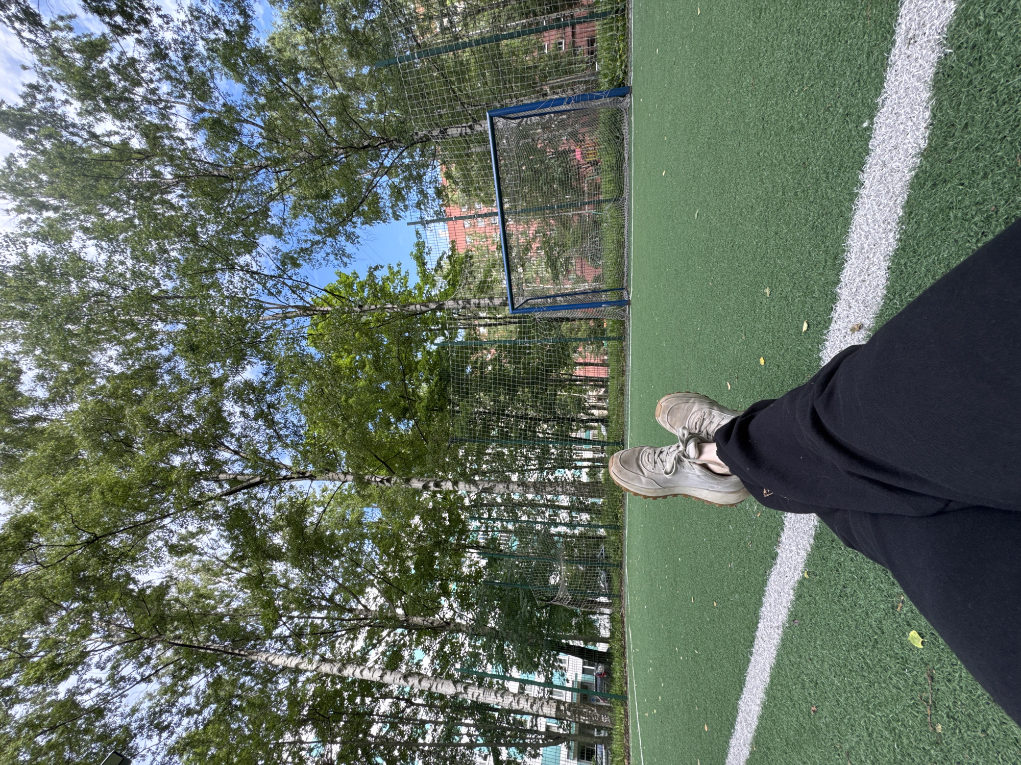 A low-angle, first-person perspective shot shows a person lying on a bright green artificial turf soccer field in Saint Petersburg, Russia. The person's legs, clad in black sweatpants and light gray sneakers, are crossed and stretched out towards the camera, with the feet positioned in the lower-left quadrant of the frame. A white boundary line runs across the field in front of the person. In the background, a standard blue soccer goal with a net stands centered, further back on the field. A tall chain-link fence encloses the field, with lush green trees and the sides of apartment buildings visible beyond it. The sky above is a bright blue with scattered white clouds. The scene is set outdoors on a clear day.