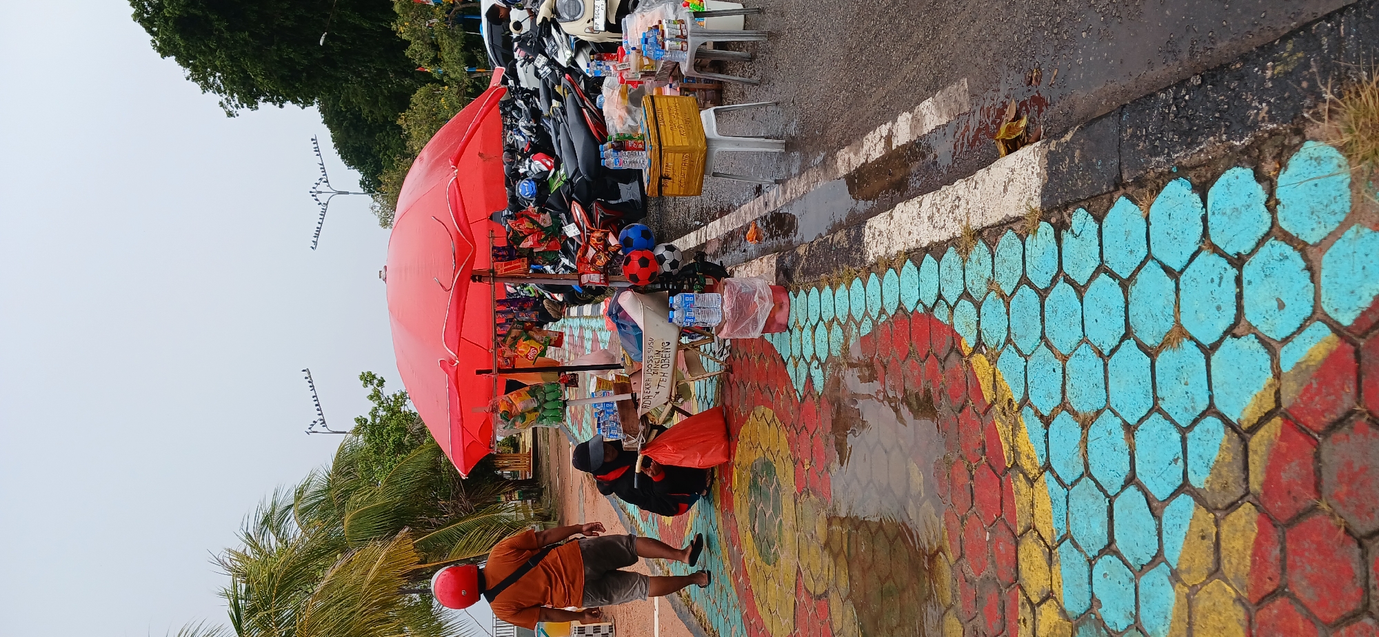 In Tanjung Balai Karimun, Indonesia, a street vendor operates a cart under a large red umbrella, selling beverages and snacks. The cart is situated on a sidewalk paved with colorful hexagonal tiles, some of which are wet and reflect the overcast sky. To the left, a person wearing an orange t-shirt and red helmet walks away from the vendor. A second person, dressed in dark clothing and a hat, kneels by the cart, handling a red plastic bag. The area is lined with parked motorcycles, suggesting a public space or thoroughfare. In the background, palm trees and dense foliage frame the scene. The ground is wet, indicating recent rain.