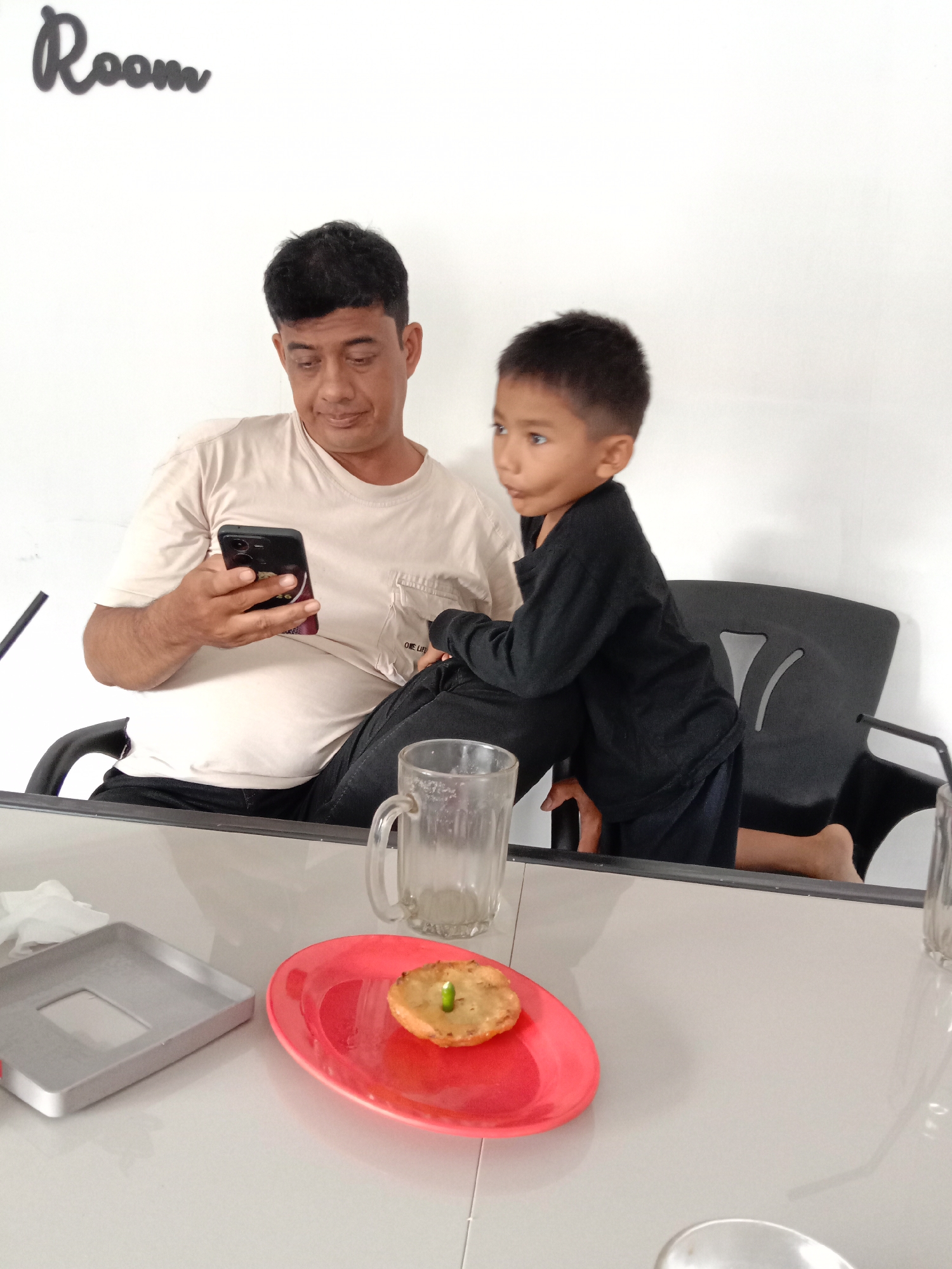 A man and a boy are seated at a table in a room, possibly in Lhokseumawe, Indonesia. The man is seated, looking at a smartphone. The boy is leaning against the man's leg. On the table, there is a glass, a plate with a small round food item topped with a green sprout, and a silver tray. The word "Room" is written on the wall in the background.