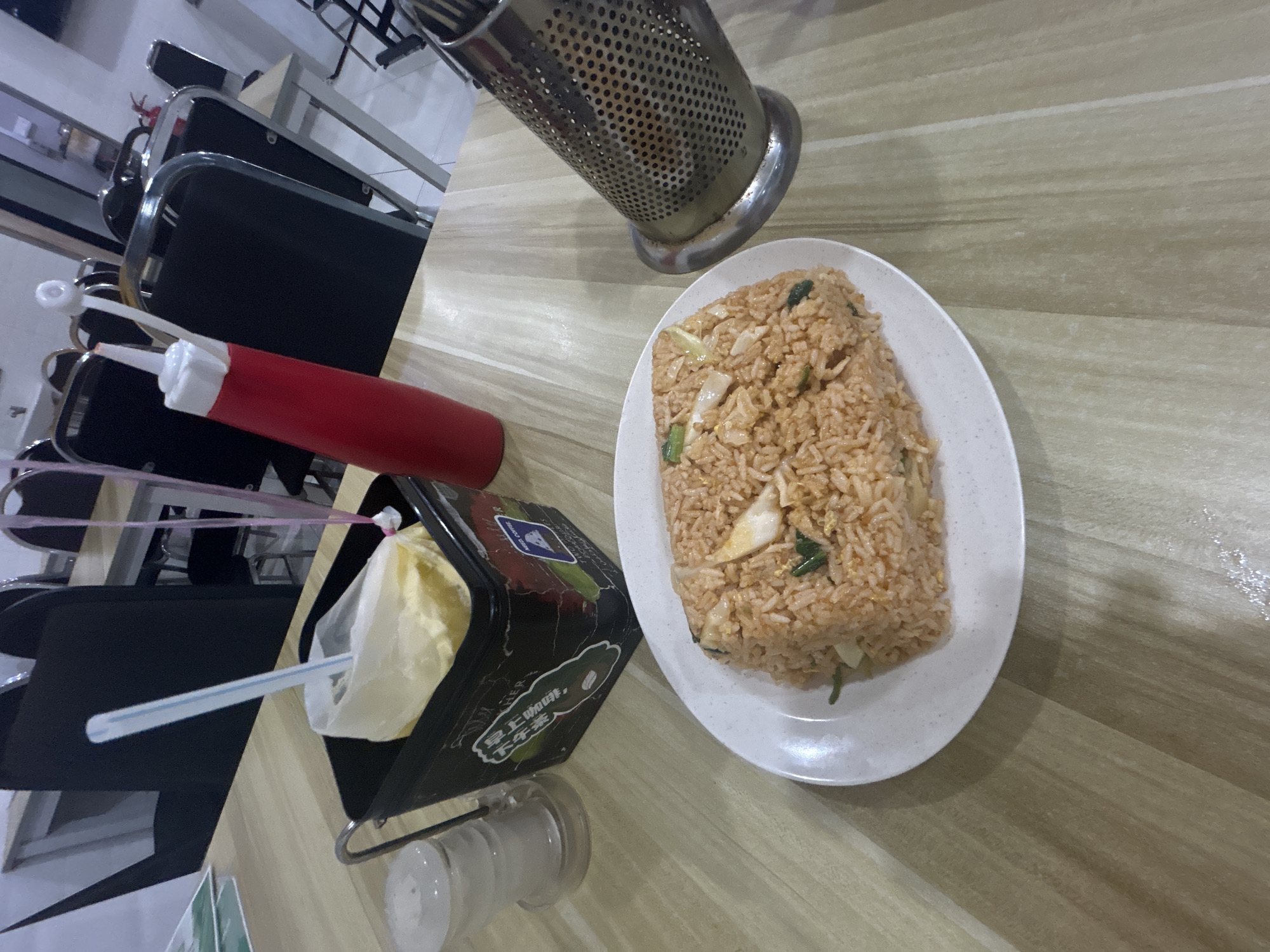 A close-up, eye-level shot shows a plate of fried rice on a wooden table in a restaurant in Kuala Lumpur, Malaysia. The fried rice, a portion of which is visible, appears to be a main dish. To the left of the plate is a black container with a plastic bag of what looks like condiments or napkins and a straw. Next to it is a clear salt shaker. In the background, a red squeeze bottle with a white top is visible, along with chairs and tables, indicating a dining area. The overall scene suggests a casual dining experience.